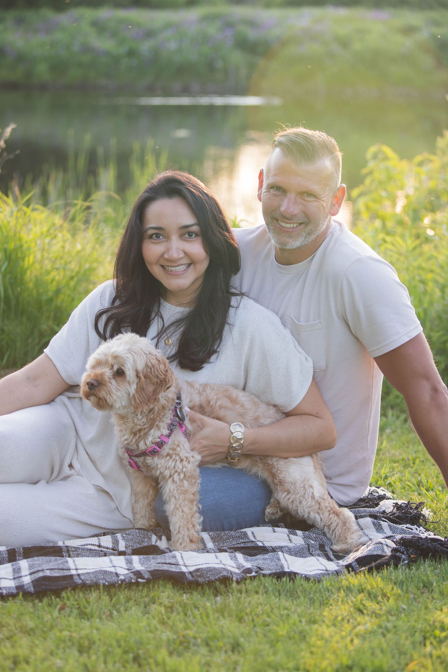 Smiling couple sitting on plaid blanket in grass with fluffy dog near pond.