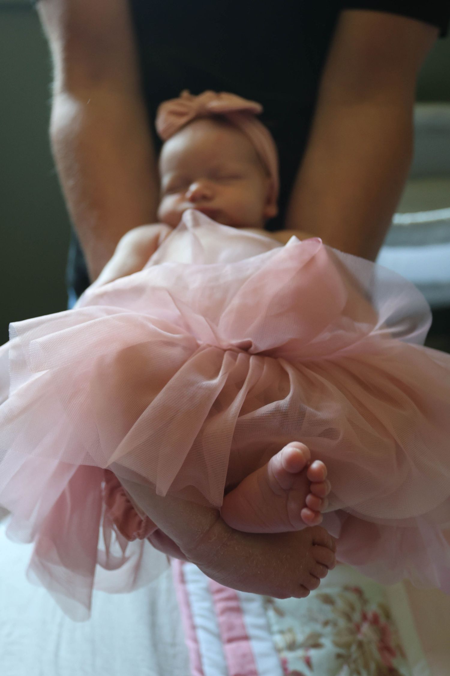Sleeping newborn baby girl in pink tutu dress with bow headband held by adult hands.
