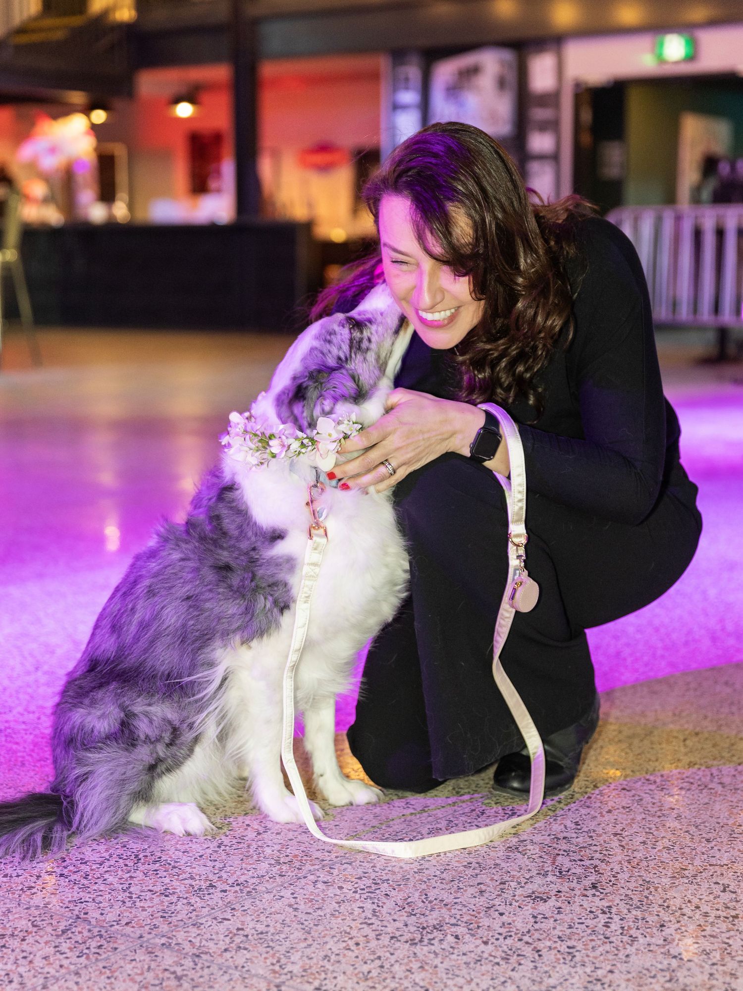 Smiling woman in black outfit being licked by adorable dog with floral crown on pink dance floor.