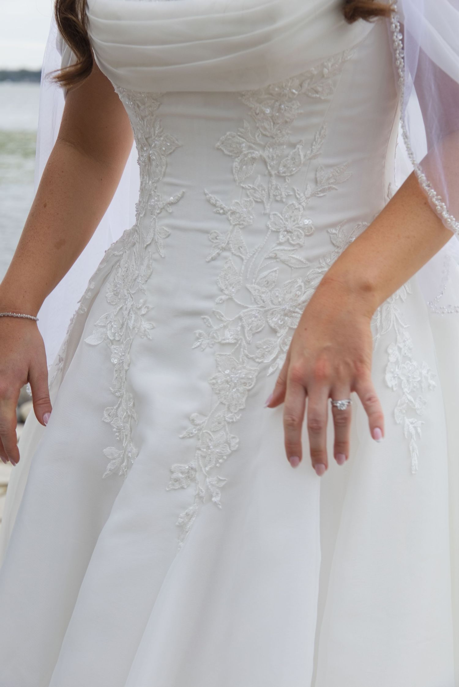 Close-up of a bride in a white lace wedding gown with floral appliques, wearing a diamond engagement ring.