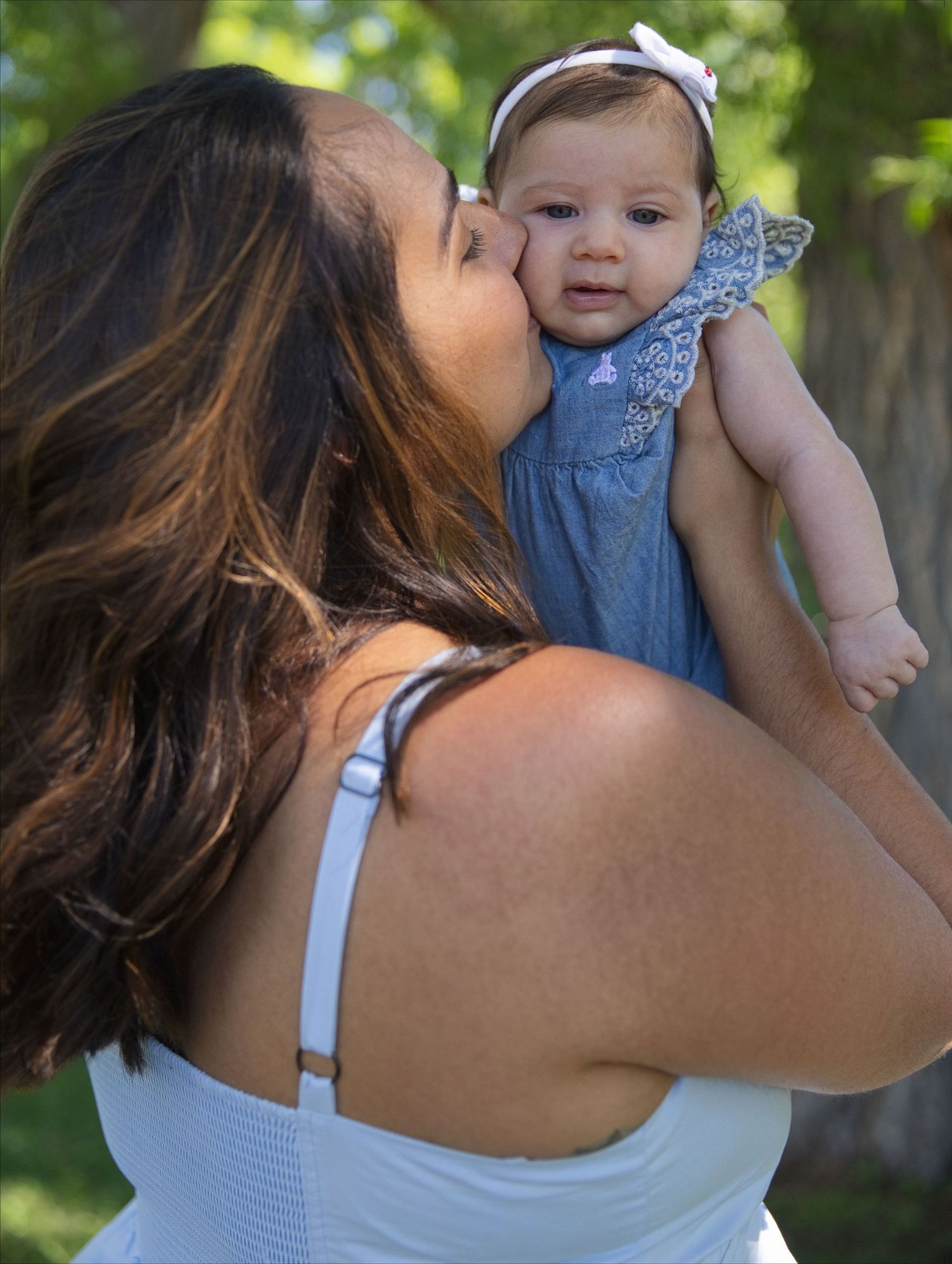 A mother in a white dress kisses her baby girl in a blue dress and white headband outdoors.