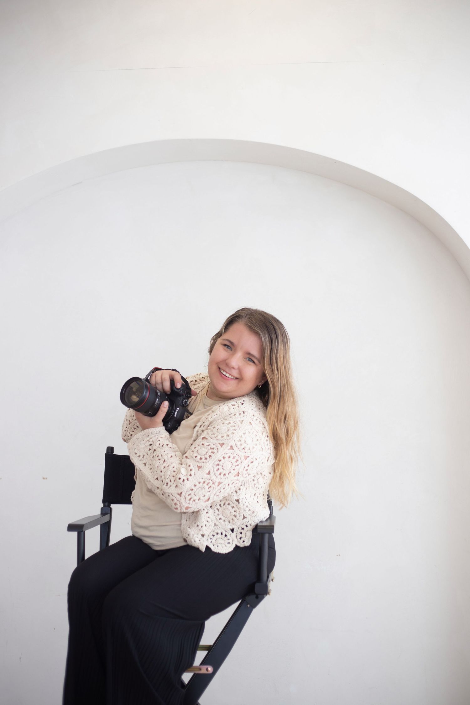 Smiling blonde woman photographer sitting in director's chair holding DSLR camera against white arched wall backdrop.