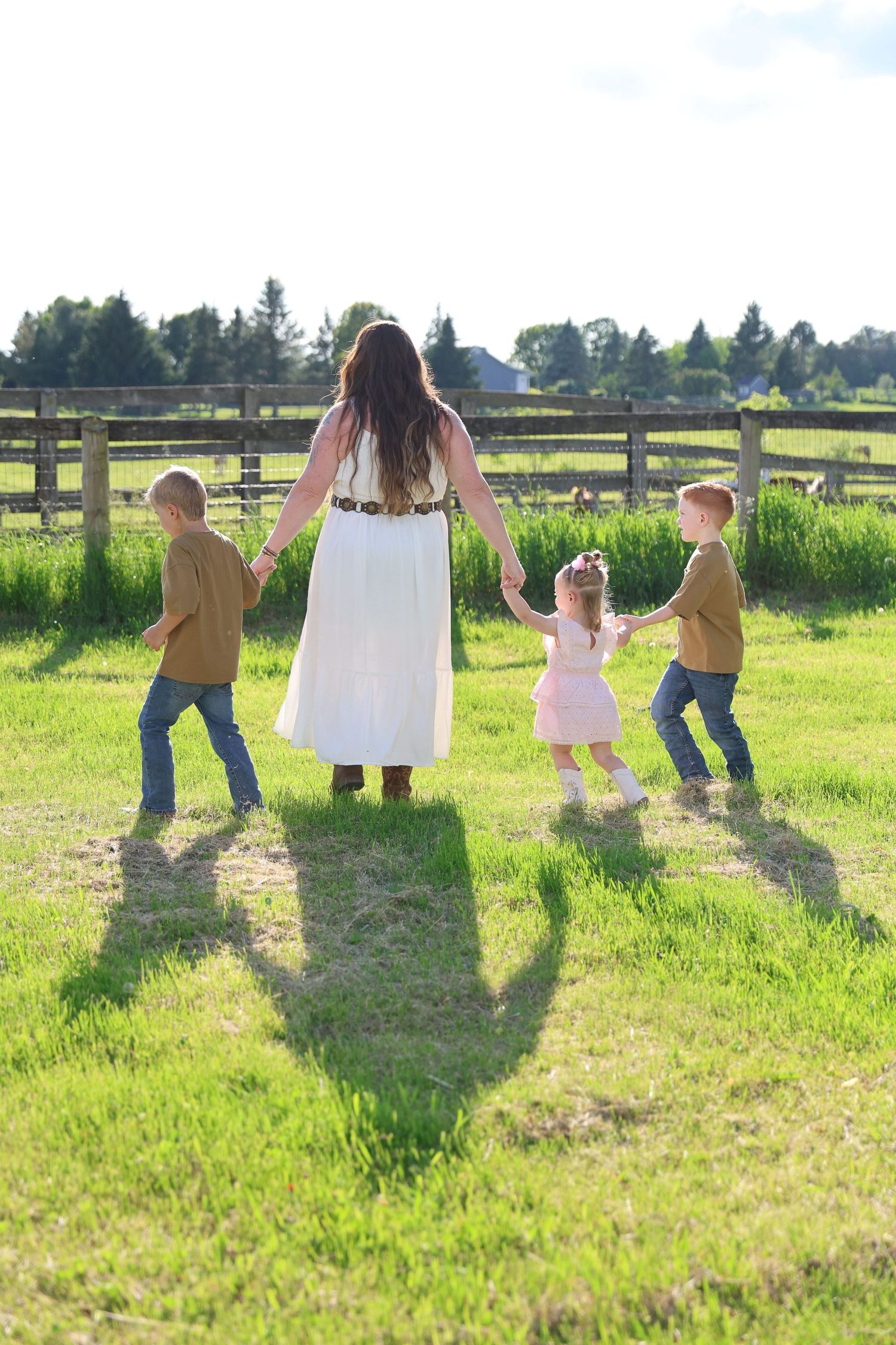 A mother in a white dress holds hands with three children while walking on green grass near a wooden fence.