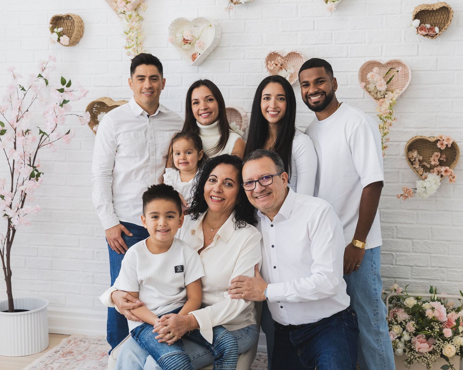 Large family portrait with eight members posing together against a white wall decorated with heart-shaped wreaths and flowers.