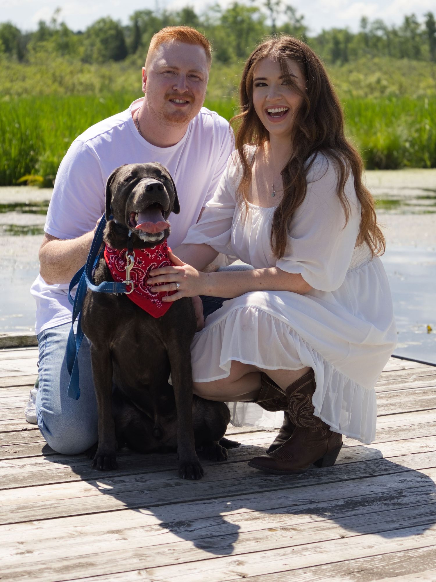 Couple smiling on a wooden dock with a black Labrador wearing a red bandana, surrounded by lush greenery and water.