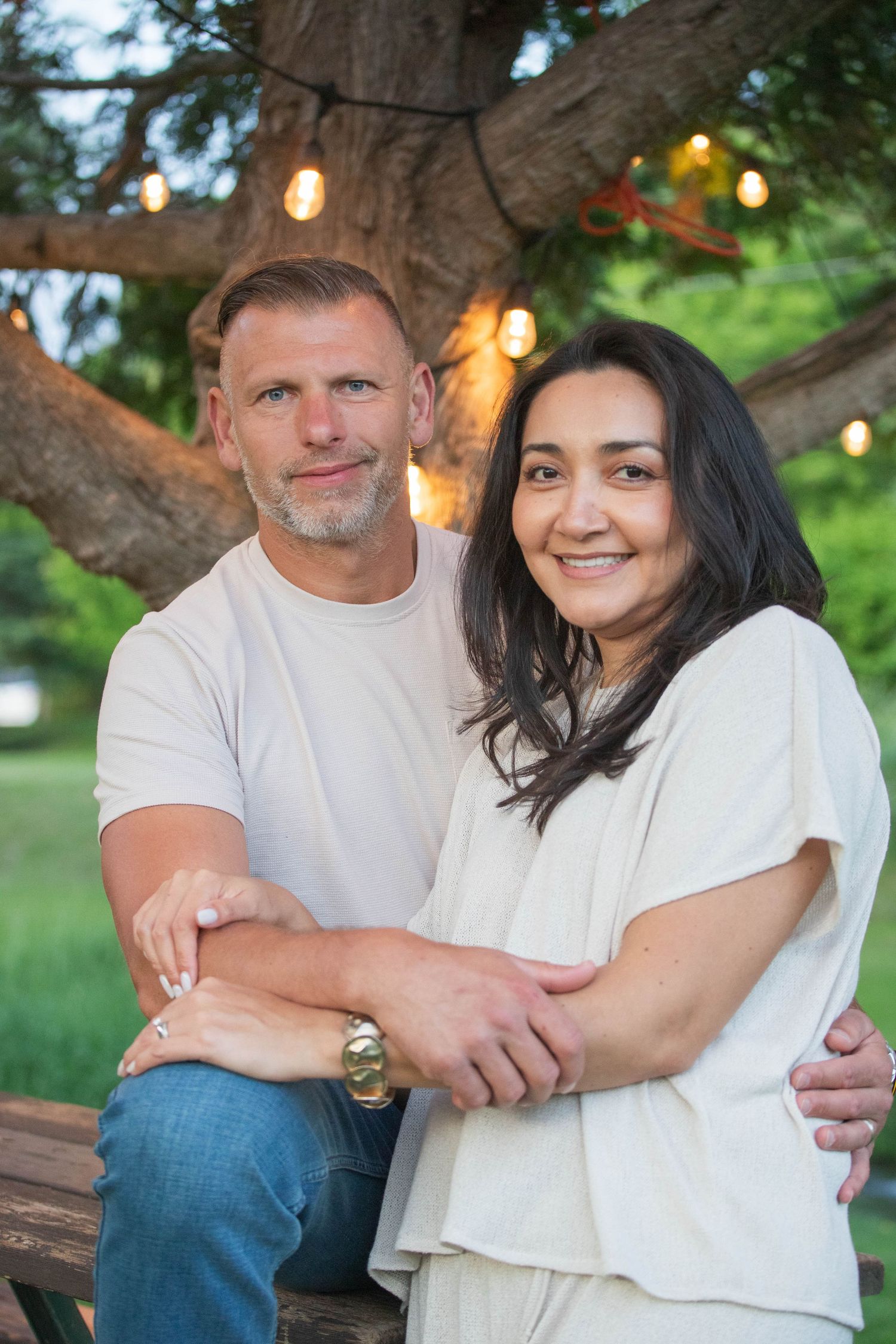Smiling couple posing together under tree with string lights in outdoor garden setting.