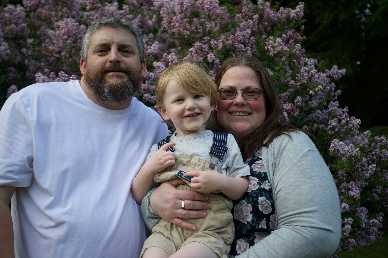 Family of three smiling in front of blooming lilac bushes.