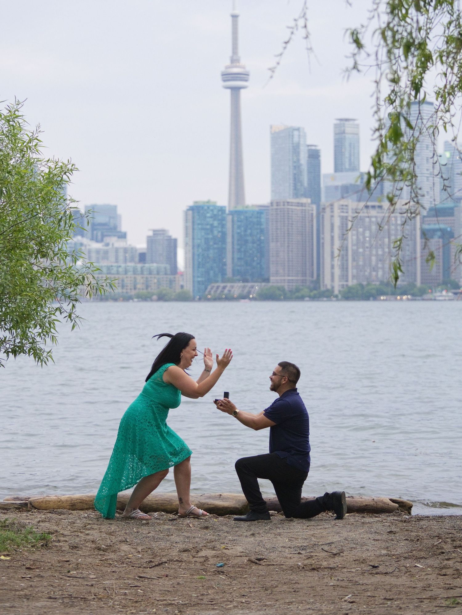 Man proposes on one knee to woman in green dress on Toronto waterfront with CN Tower skyline backdrop.