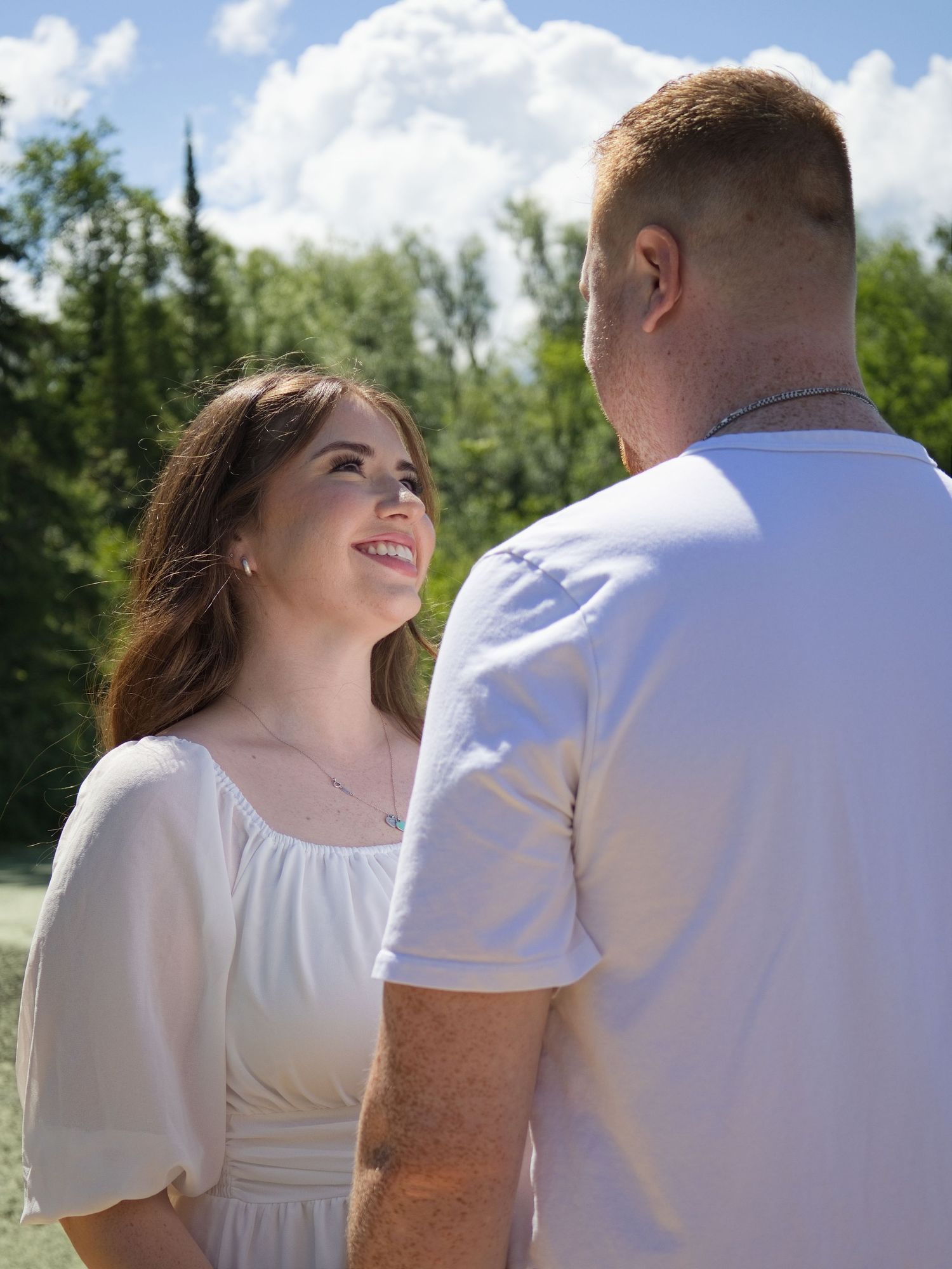 A smiling woman in a white dress gazes at a man in a white t-shirt outdoors on a sunny day.