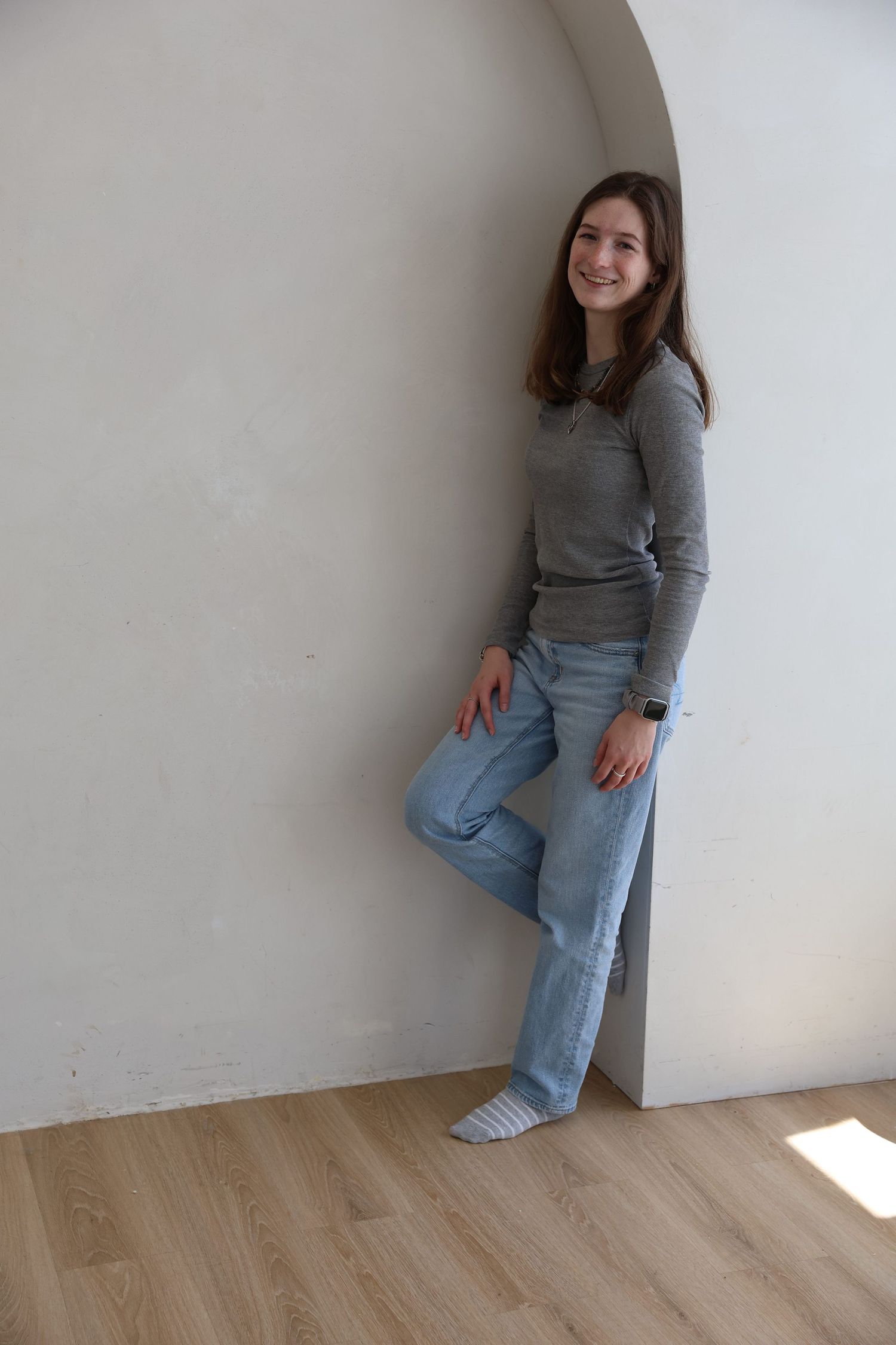 Young woman in gray sweater and light blue jeans leaning against white wall in arched alcove on wood floor.