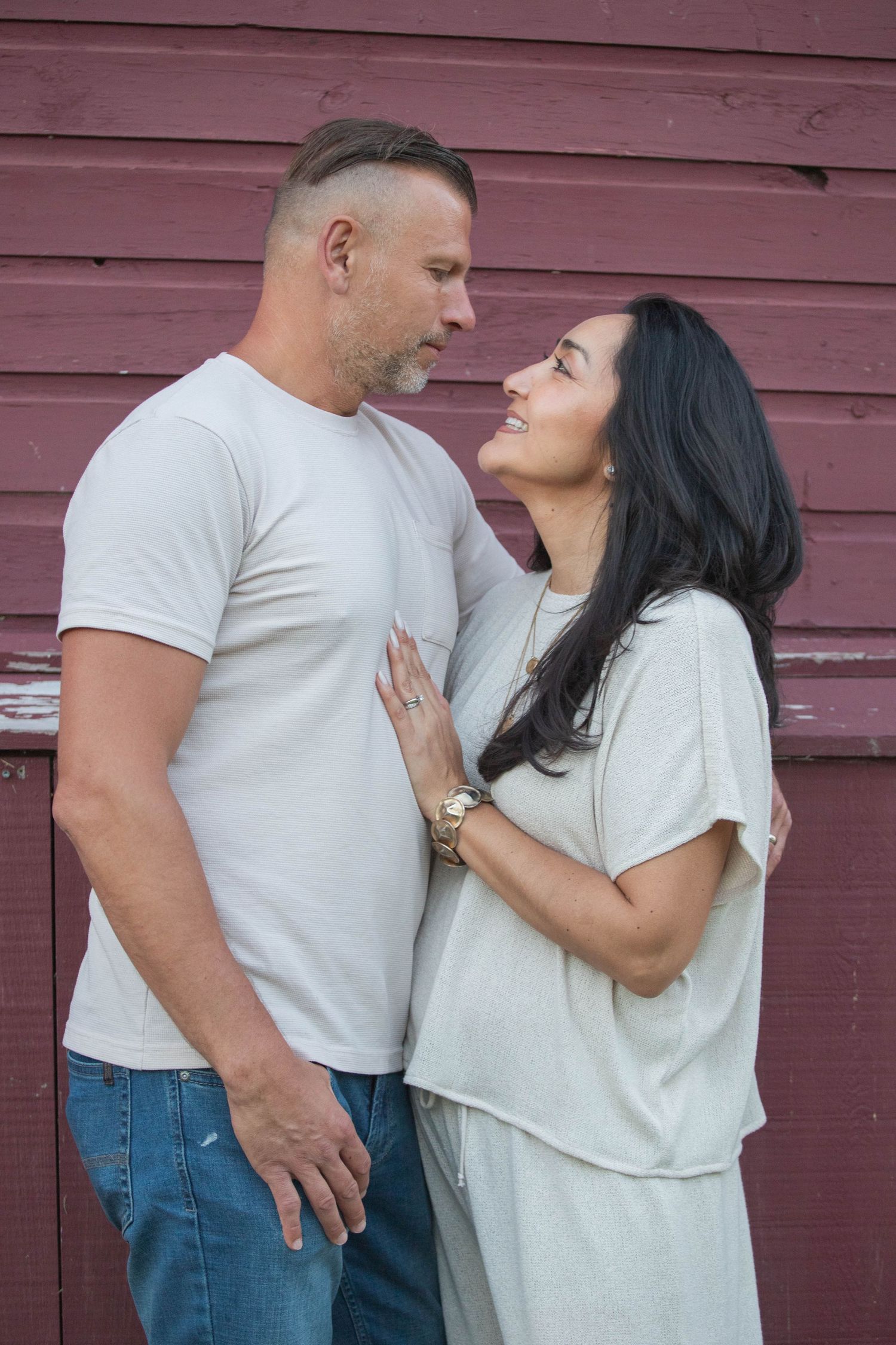 A couple in neutral tones gazes lovingly at each other against a rustic red barn wall.