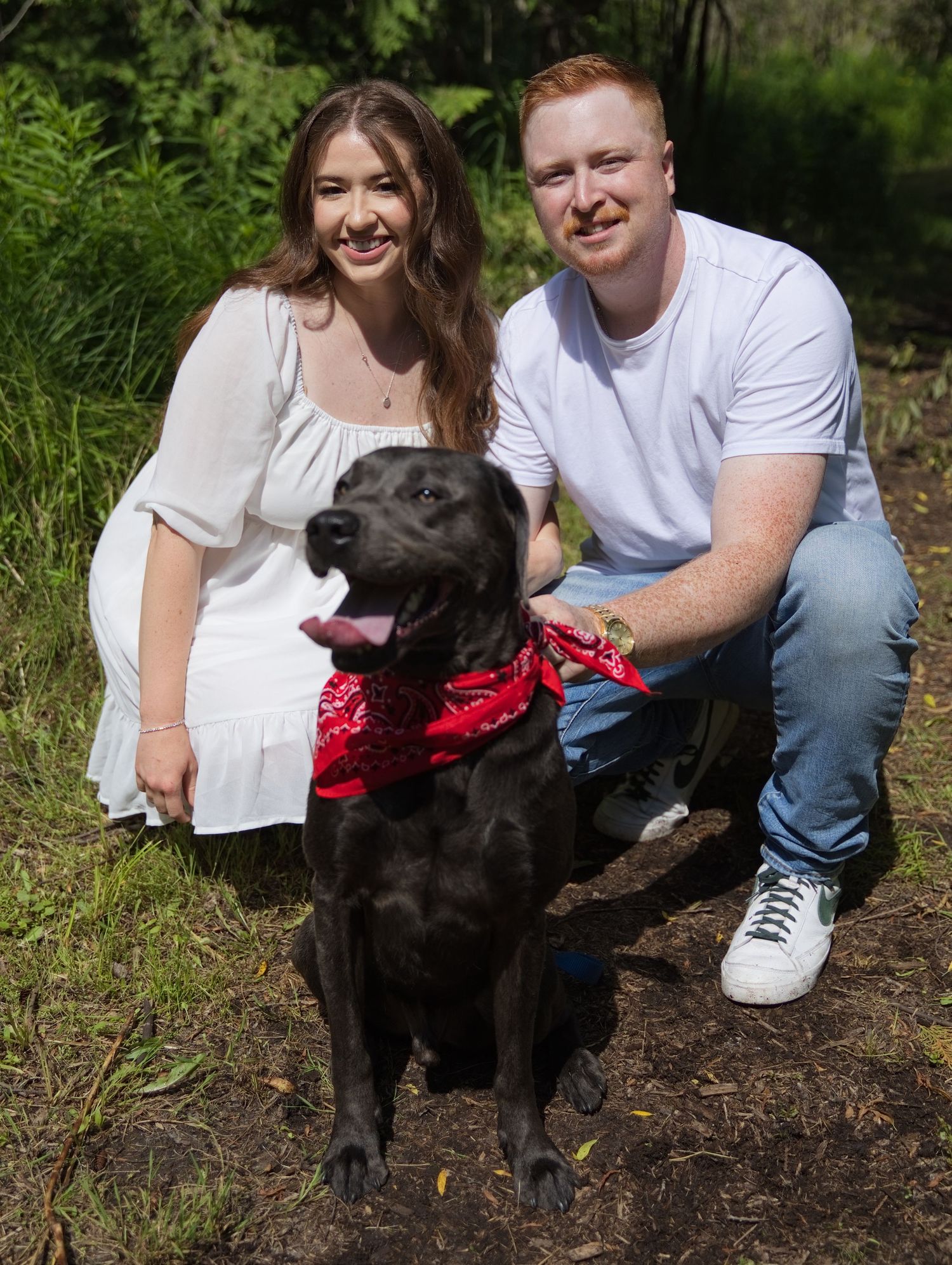 A smiling couple in casual attire poses outdoors with their black Labrador wearing a red bandana.