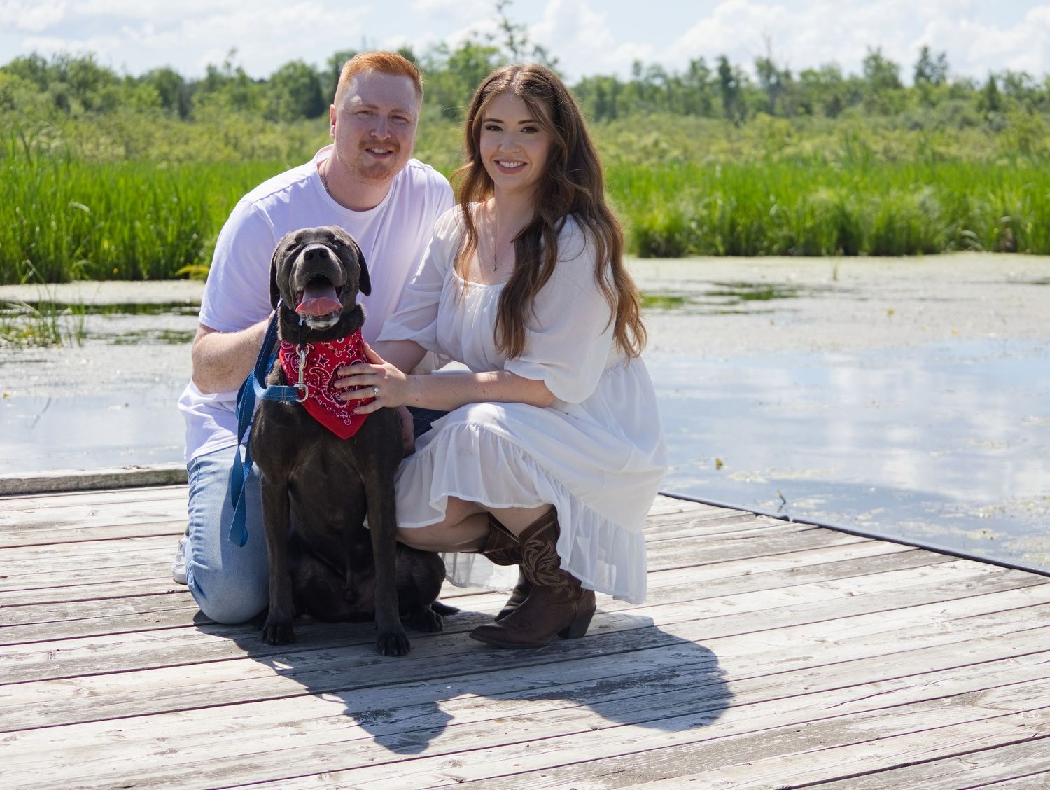 Couple with black Labrador dog posing on wooden dock near lake surrounded by green marshland.