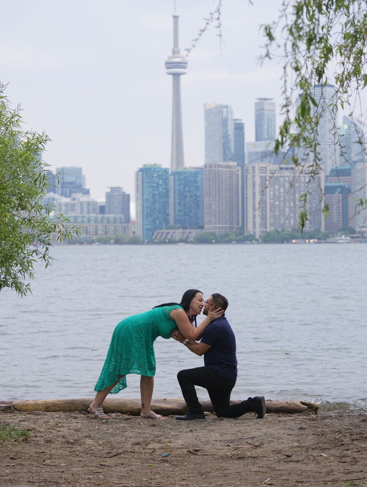 Couple sharing a romantic proposal kiss on Toronto waterfront beach with CN Tower skyline backdrop.