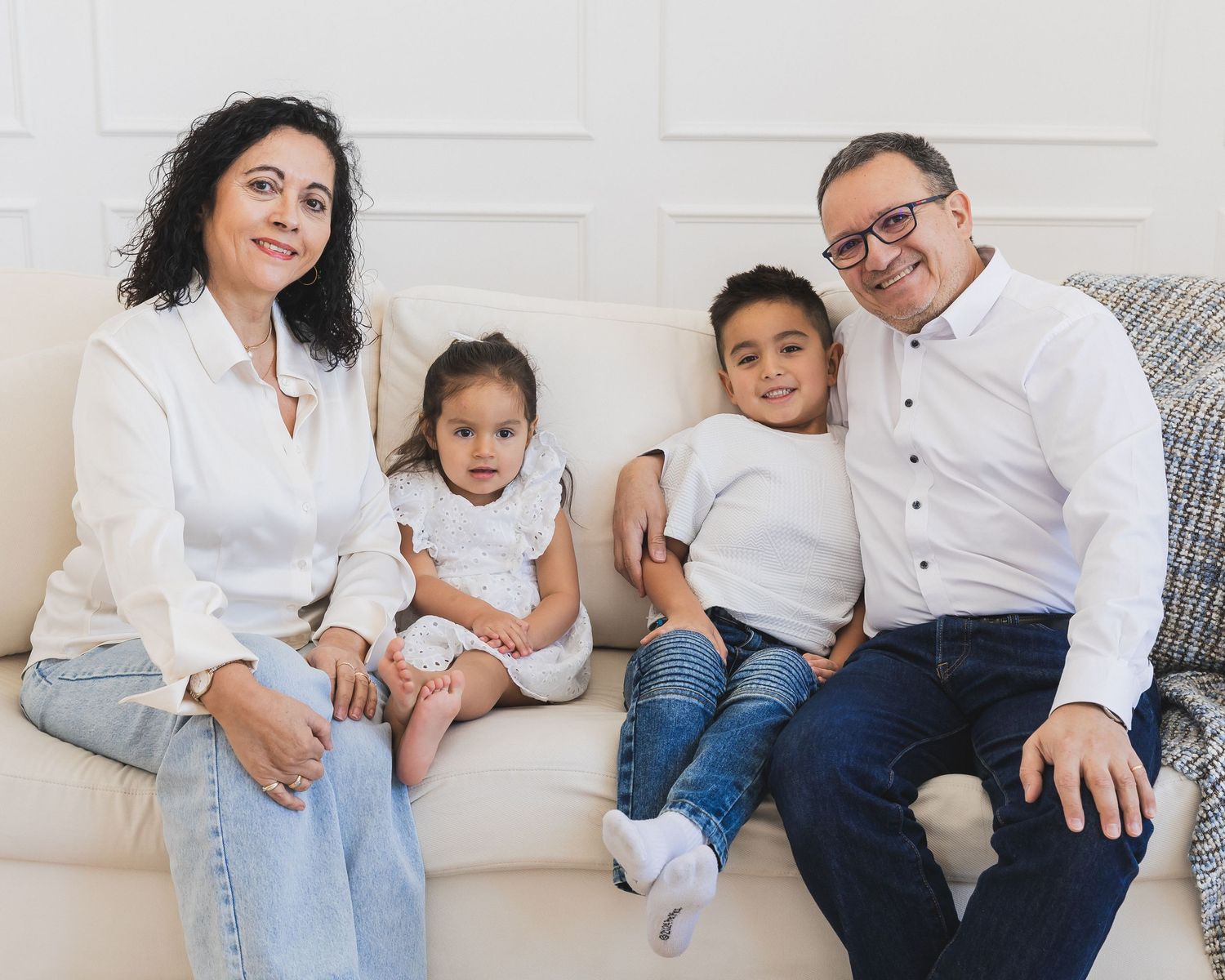 Family of four posing on a white sofa, parents sitting with two young children in a bright, elegant setting.