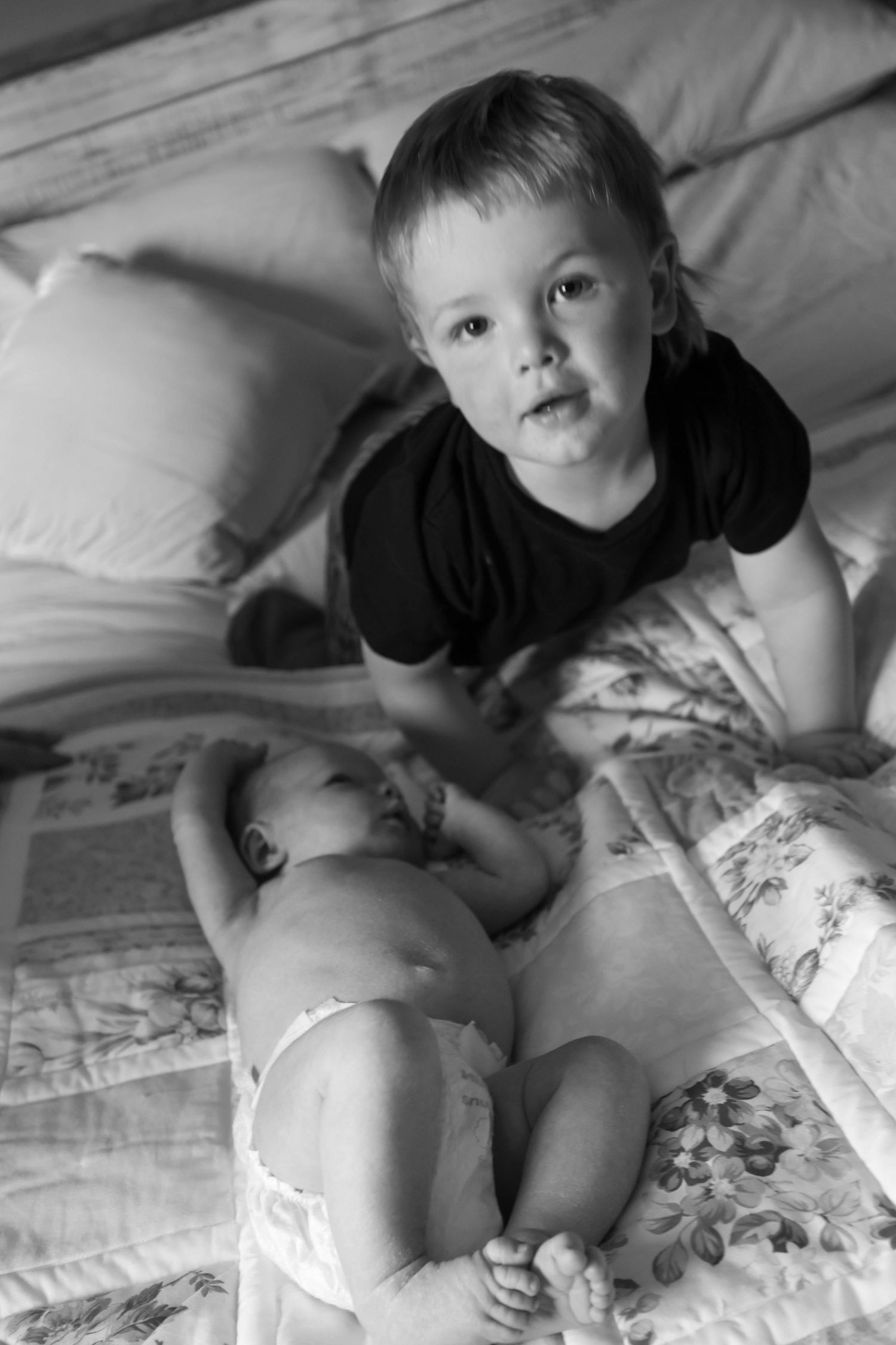 A young boy sits on a bed next to a newborn baby in black and white.