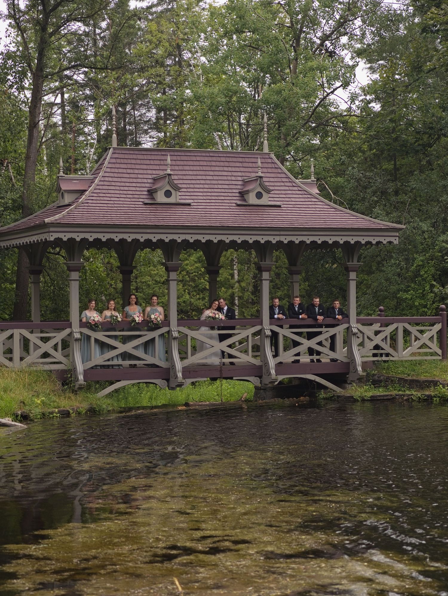 Wedding party gathered in a charming gazebo over a pond surrounded by lush green trees.