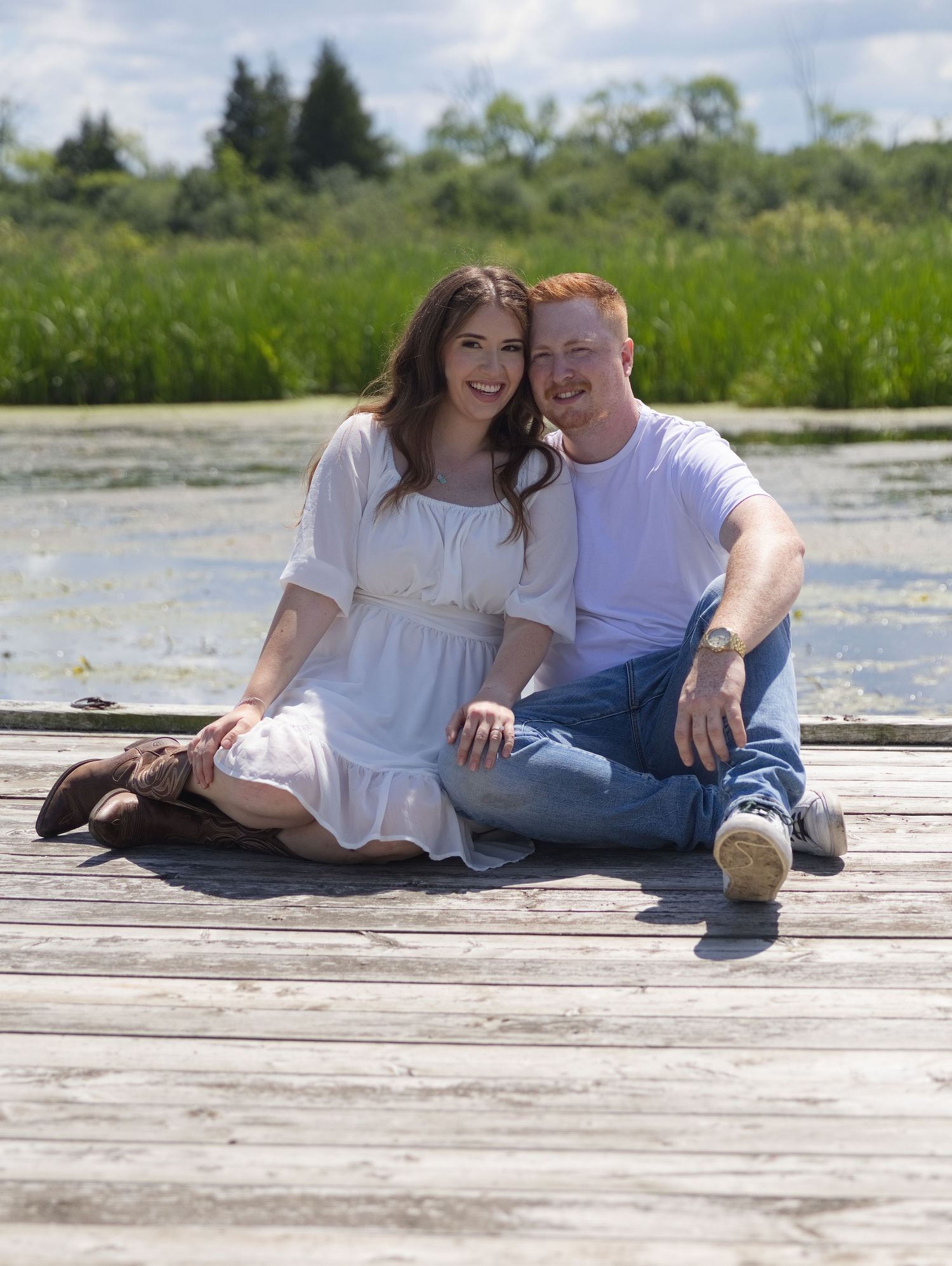Couple sitting together on a wooden dock by a serene pond, smiling, woman in white dress, man in jeans and white shirt.