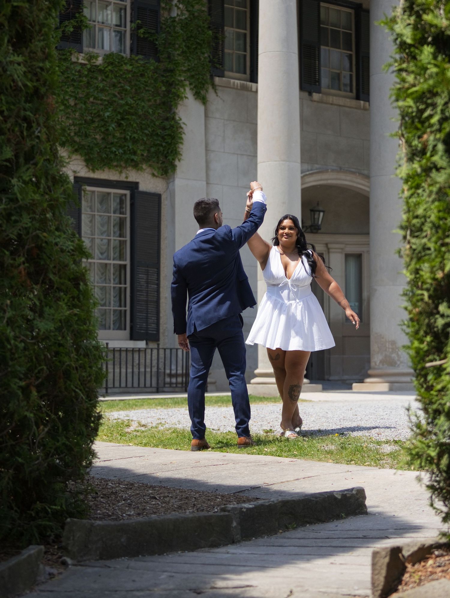 Couple dancing outdoors near ivy-covered mansion, man in navy suit spinning woman in white mini dress on sunny day.