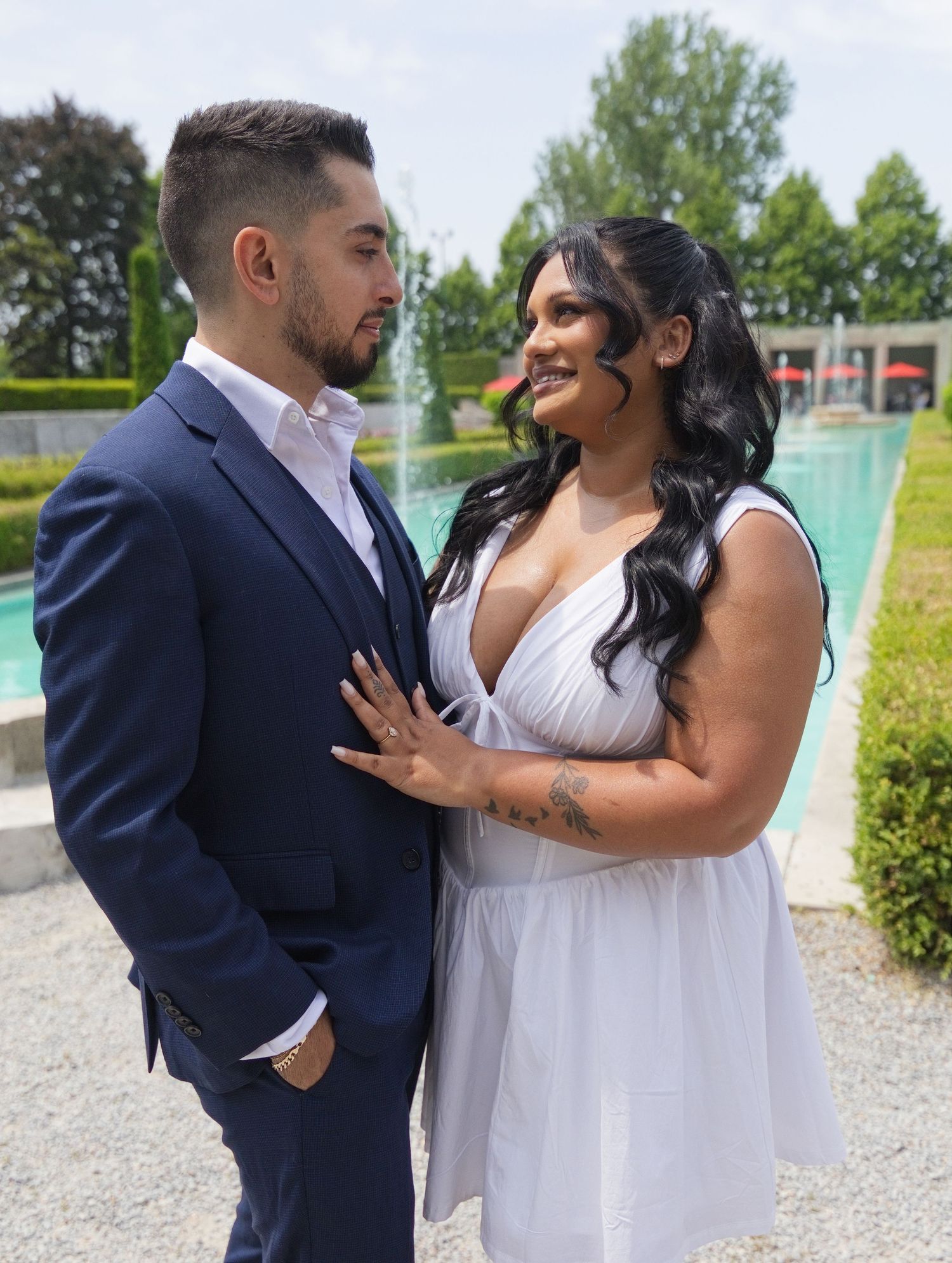 Couple in formal attire posing by a fountain outdoors, man in navy suit and woman in white dress smiling at each other.