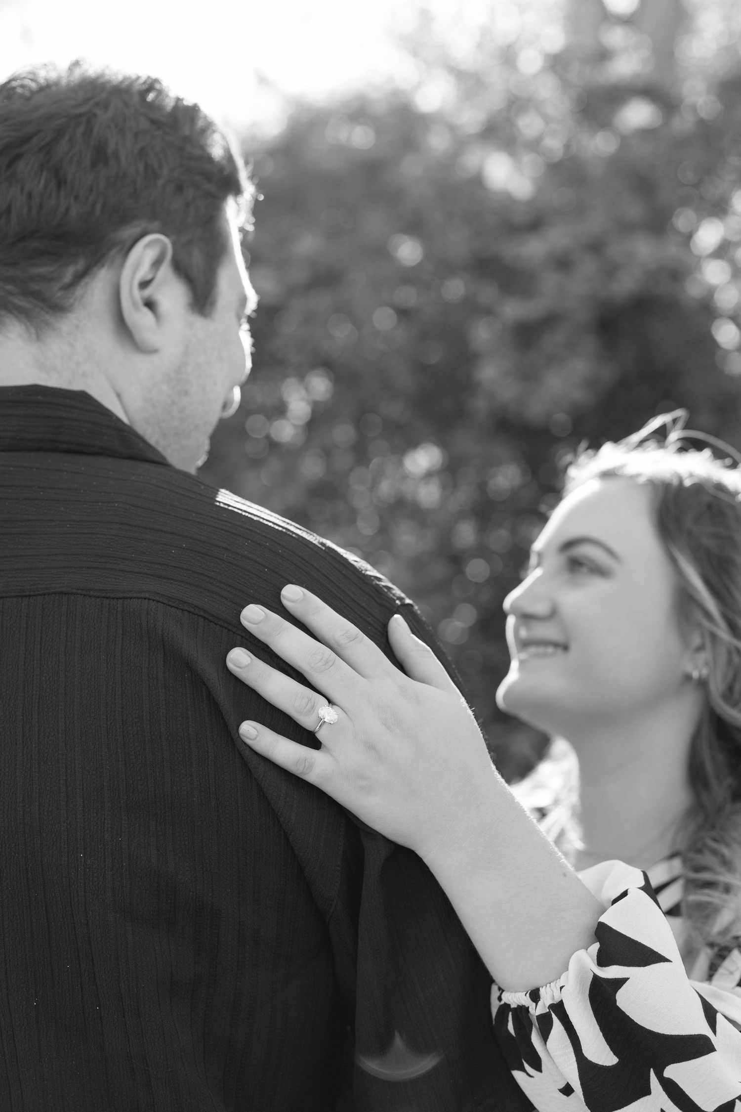 Black and white romantic photo of smiling woman with engagement ring embracing man outdoors.