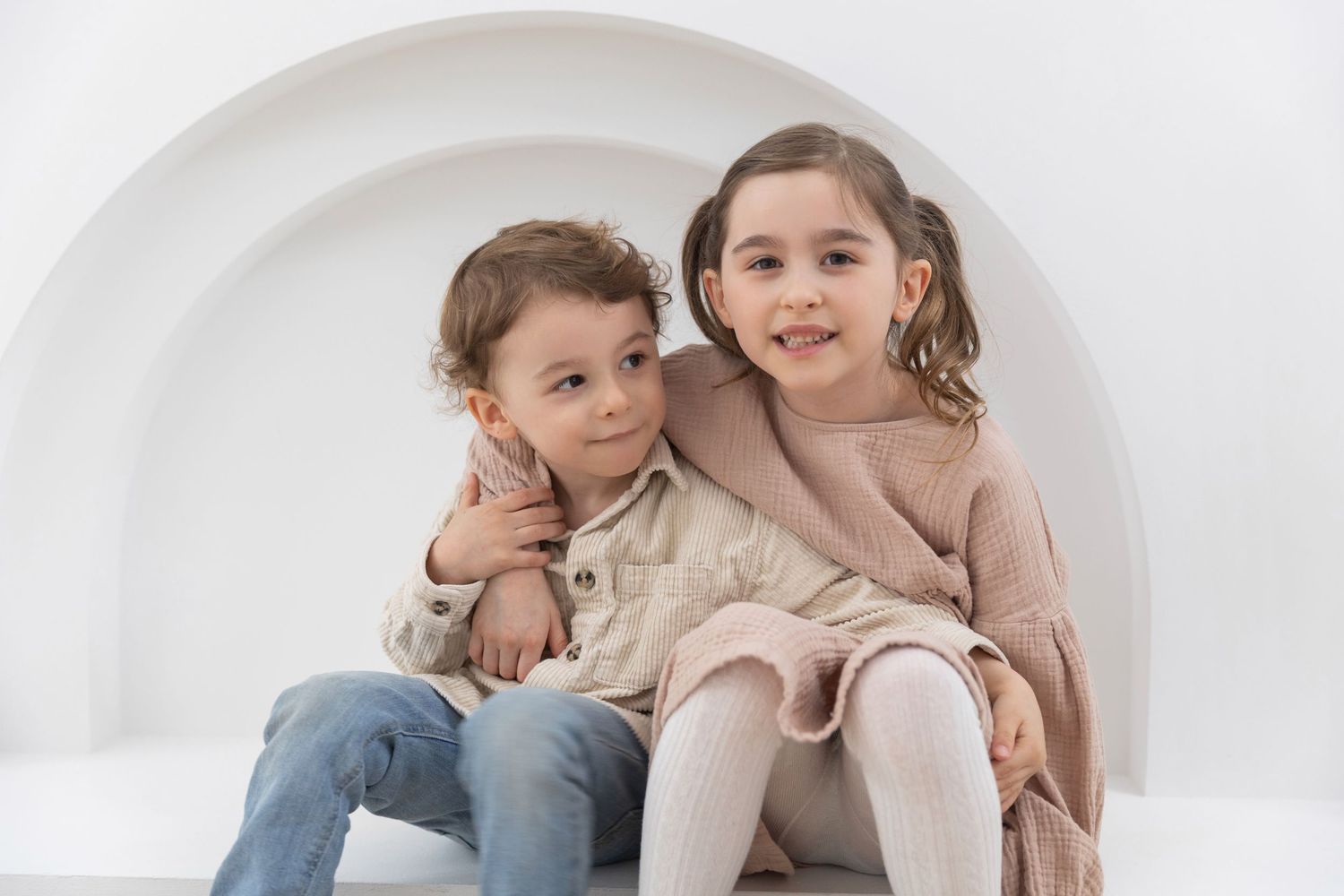Two young siblings posing together in a white studio, boy in jeans and girl in pink dress.