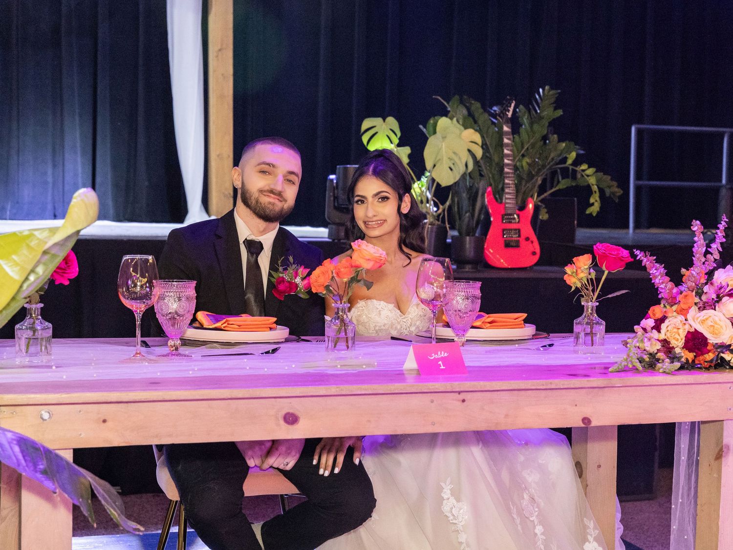 Bride and groom sit together at rustic wooden sweetheart table with guitar decor in background.