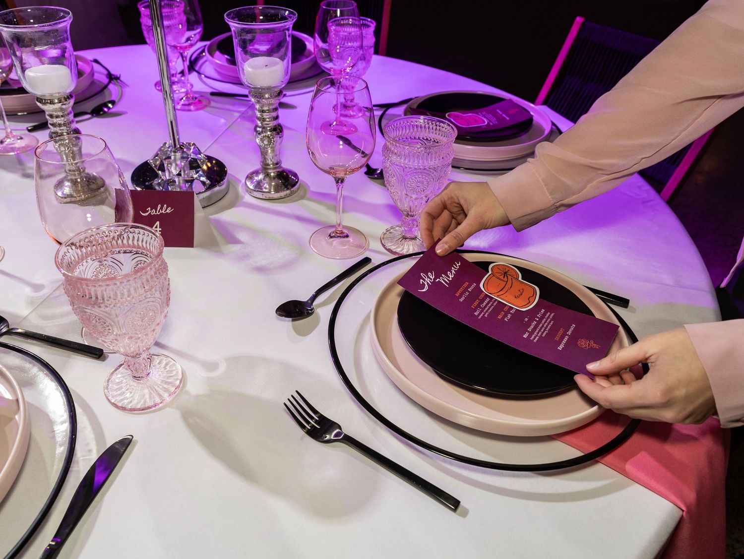 Wedding table setting with pink glassware, black plates, and a purple menu card under violet lighting.