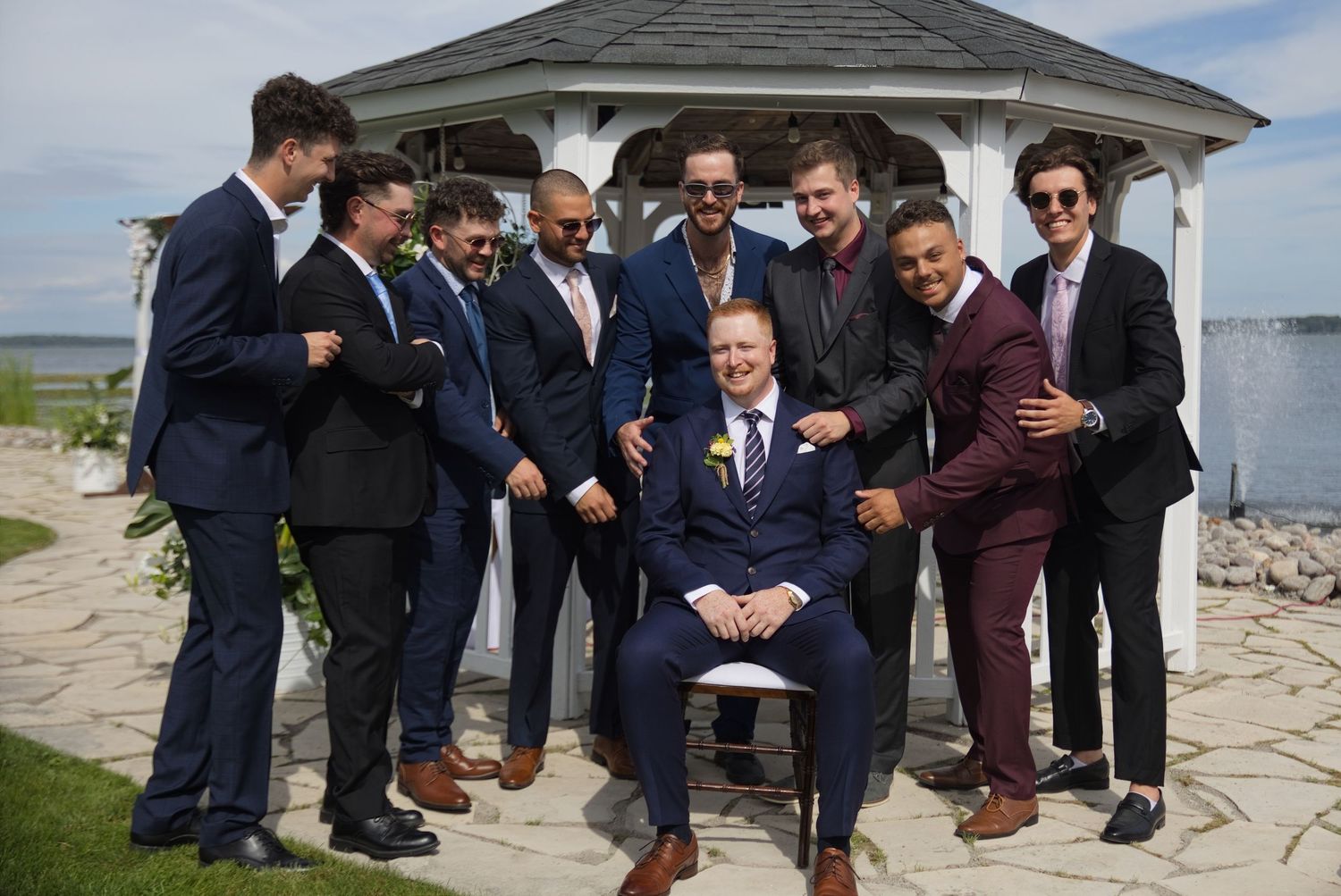 Groom seated in chair surrounded by groomsmen in navy and burgundy suits posing near a gazebo by the water.