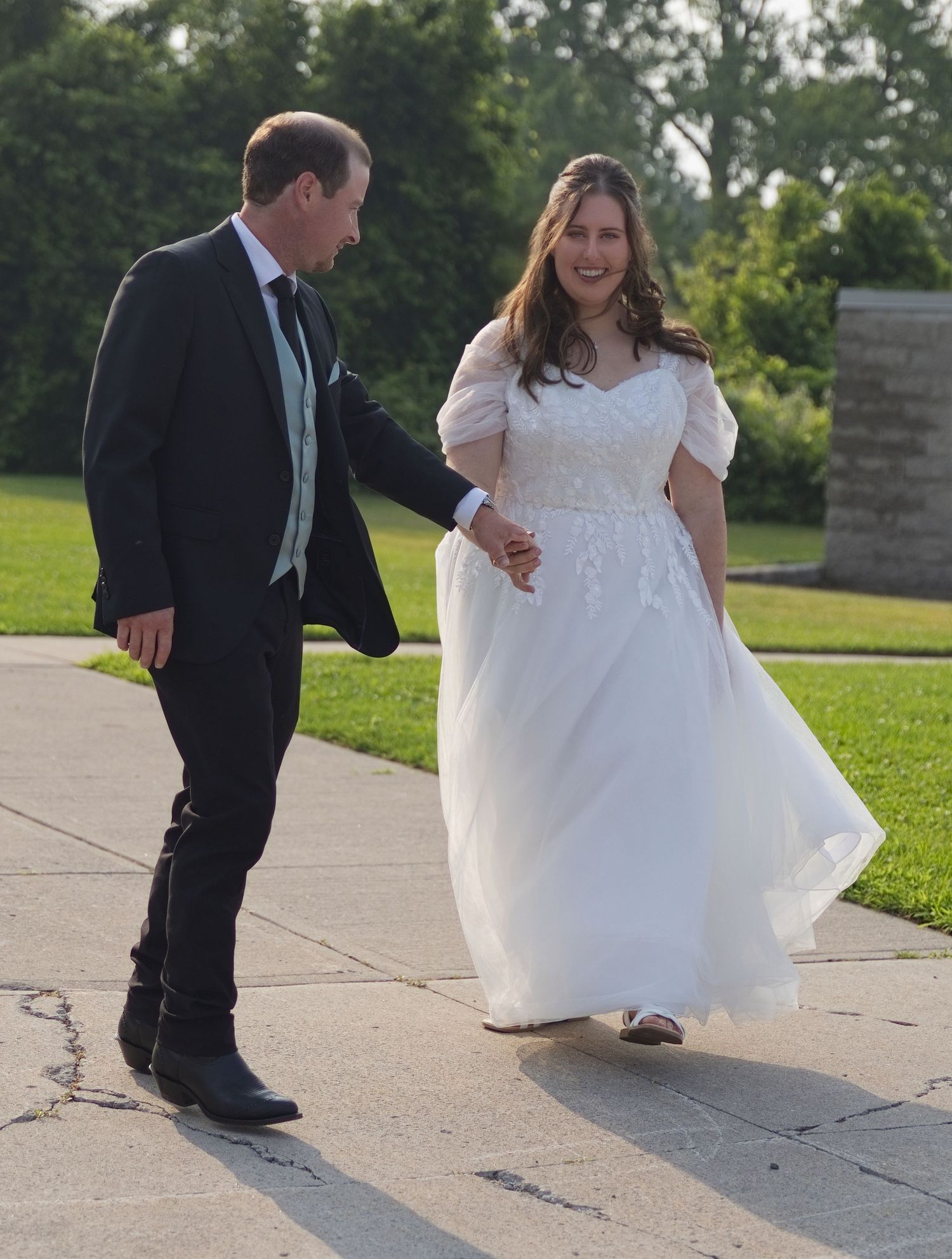 Bride in white lace gown and groom in black suit walk hand in hand on a sunny outdoor path.