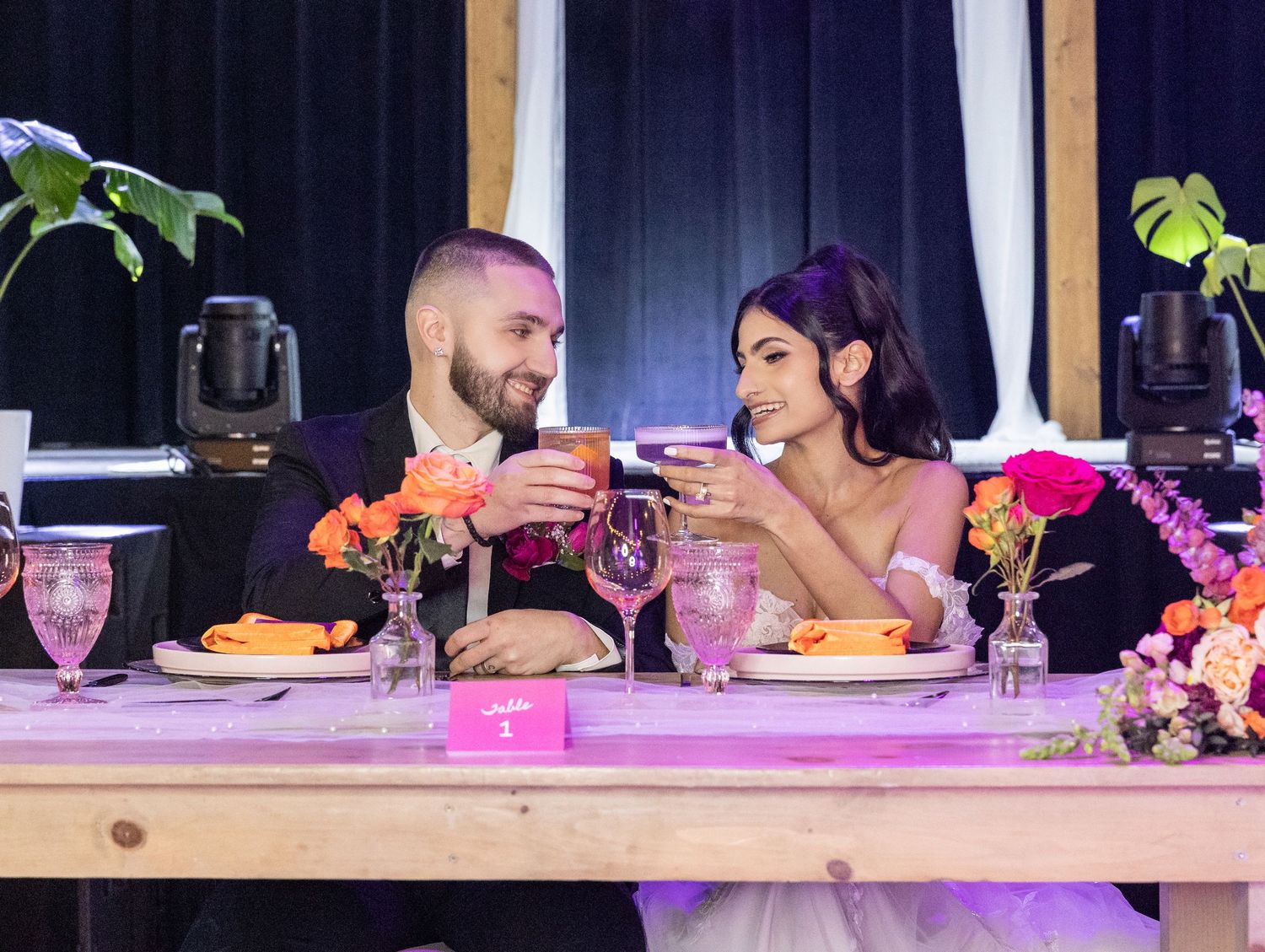 A bride and groom toast at a decorated sweetheart table with colorful floral arrangements at their reception.