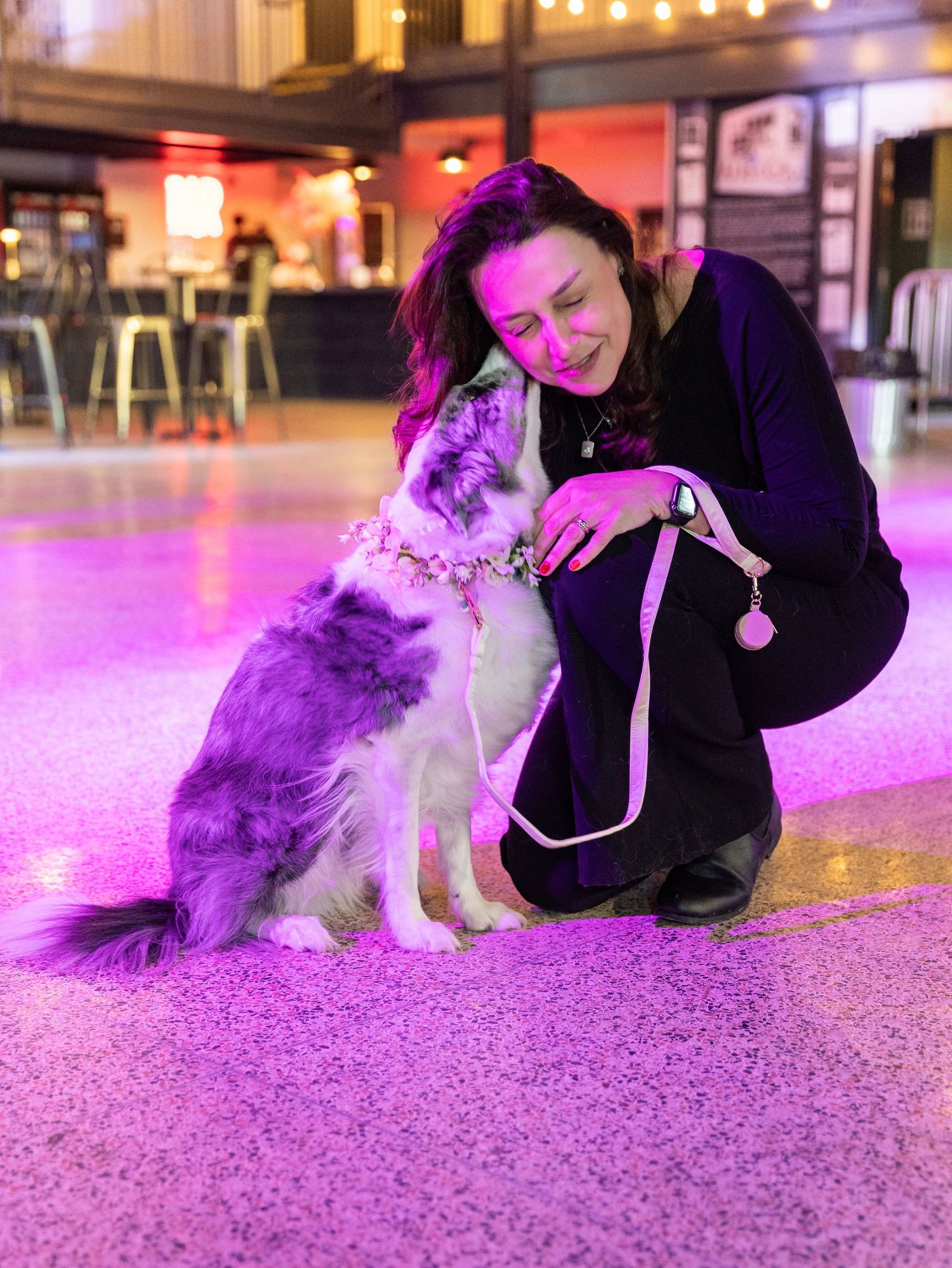 Woman in black outfit crouching with fluffy gray and white dog wearing floral crown on pink lit floor.