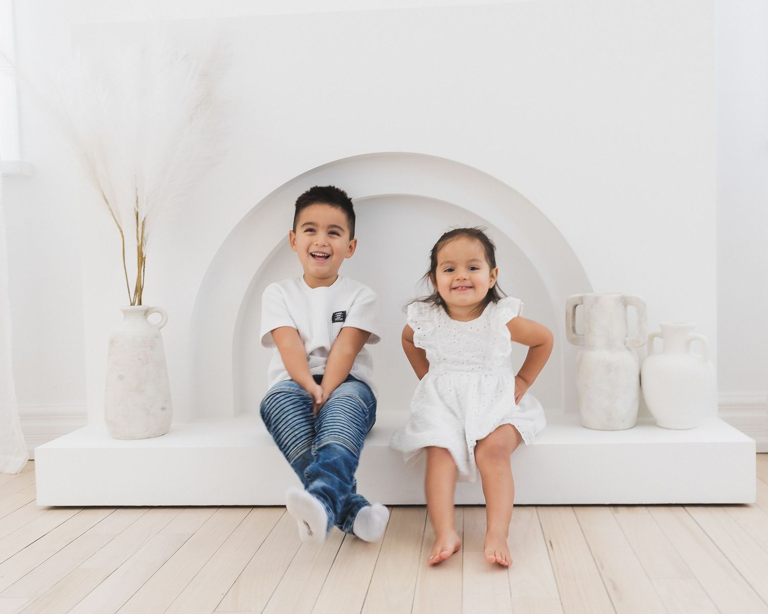 Two young siblings in white outfits sitting on bench in bright minimalist white studio.