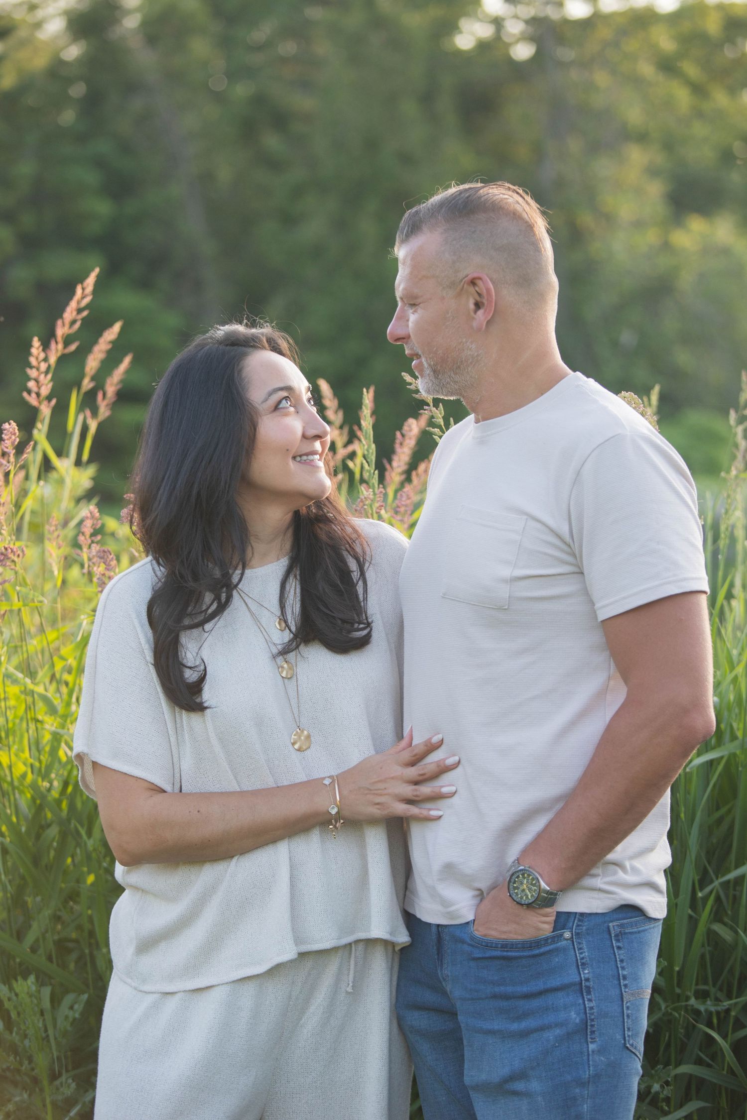 A smiling couple stands together in a sunlit field, the woman in gray and man in white tee, sharing a warm loving gaze.