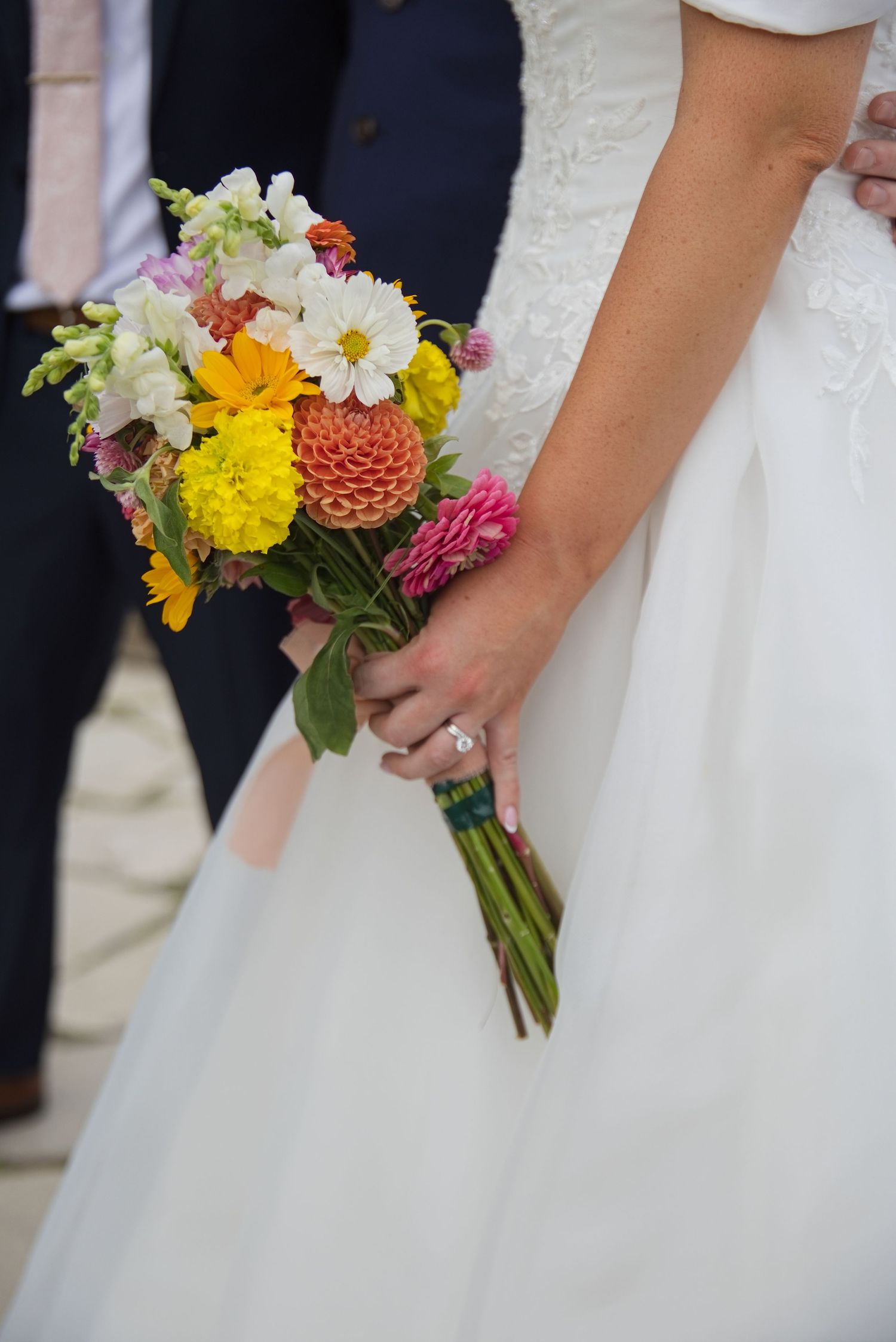 Bride holding vibrant colorful wedding bouquet with dahlias and daisies in white lace gown.