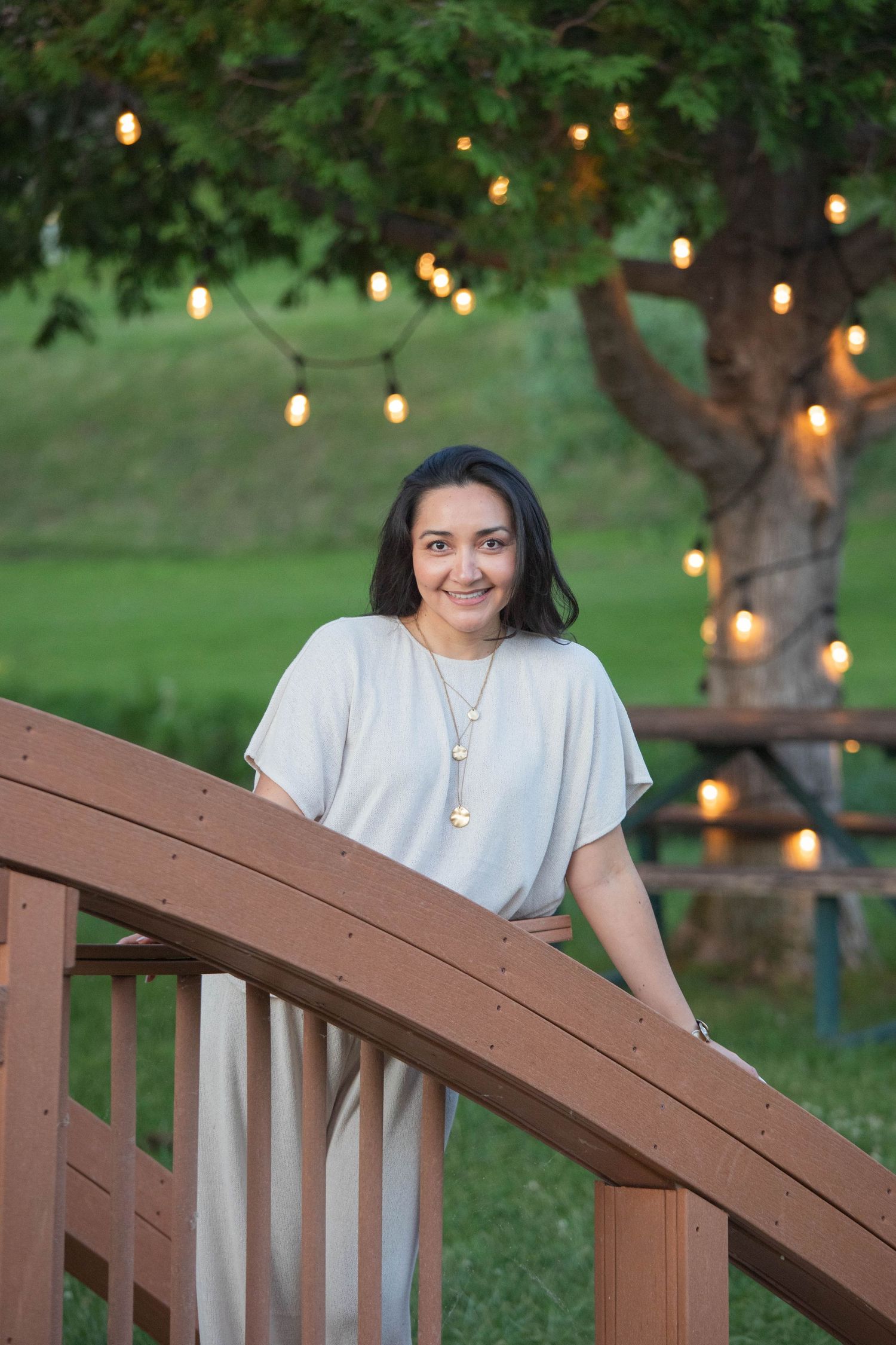 Smiling woman leaning on wooden bridge railing with string lights in tree.