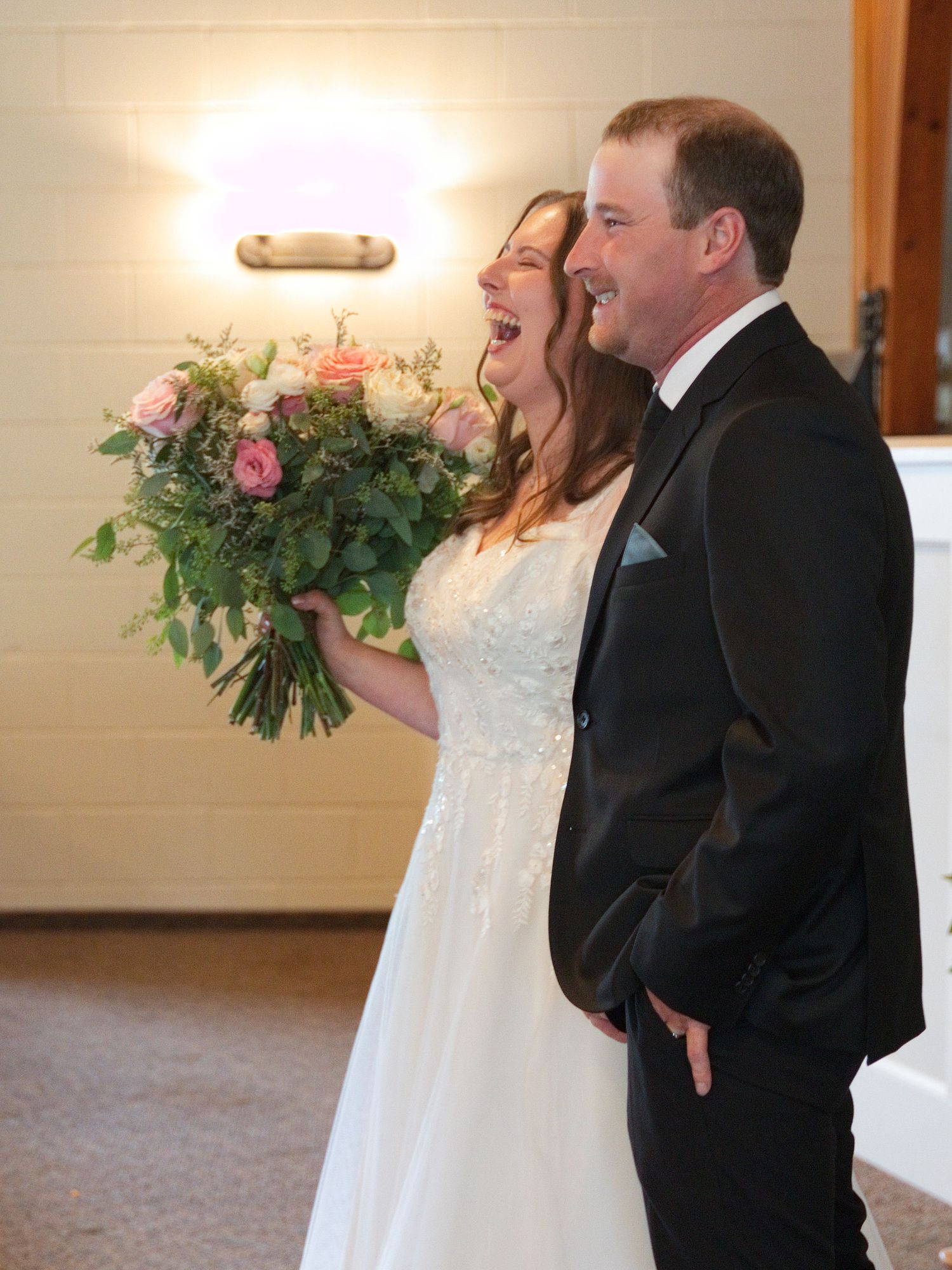 Joyful bride holding pink rose bouquet laughing with groom in black tuxedo at elegant wedding ceremony.
