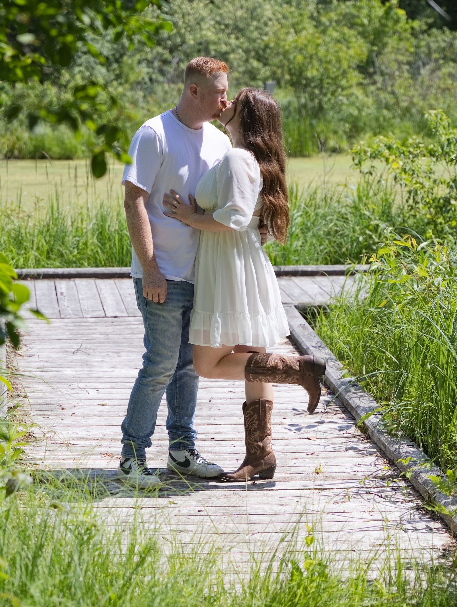 Couple sharing a romantic kiss on a wooden bridge surrounded by lush green grass and trees on a sunny day.