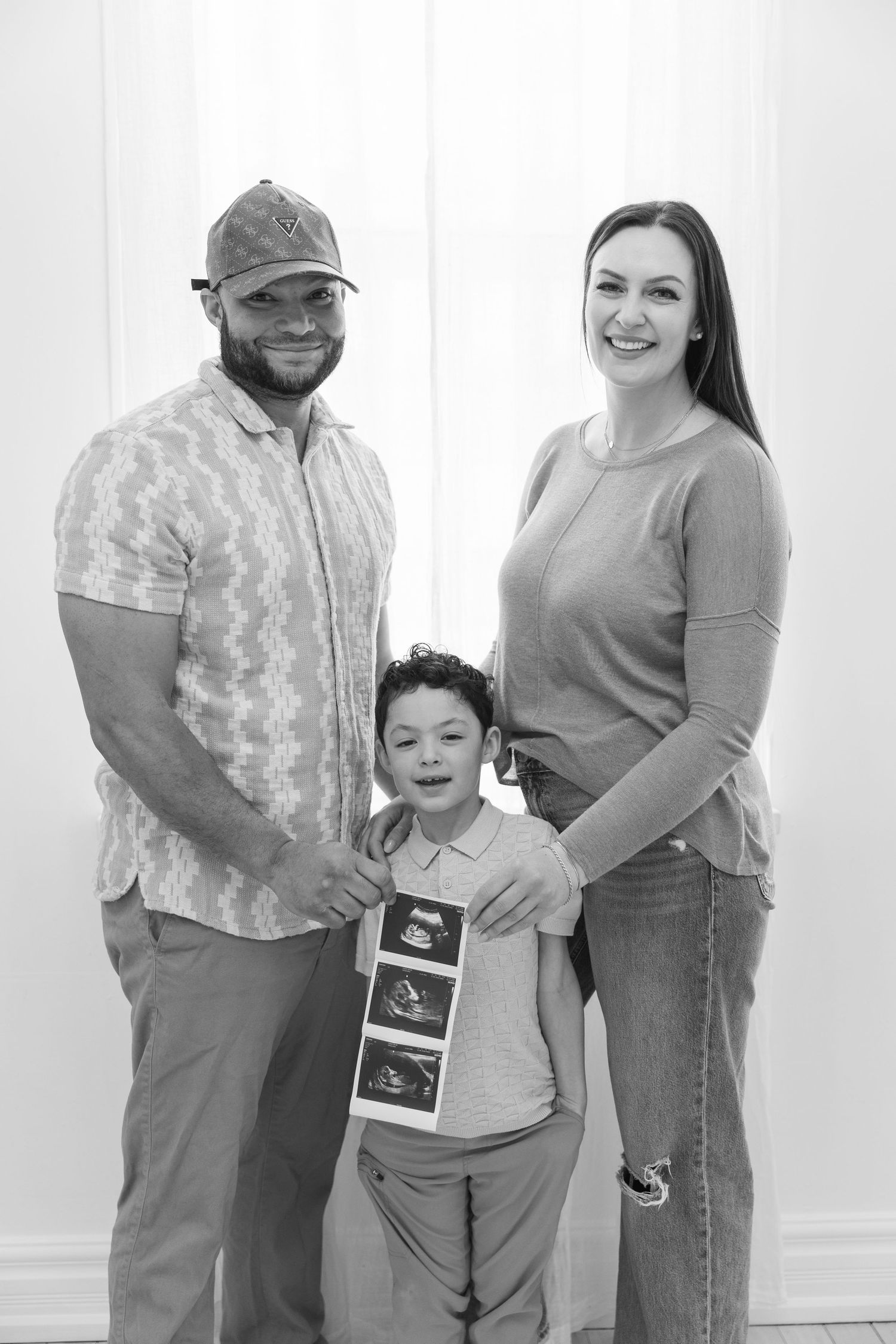 Black and white family photo with parents and young boy holding ultrasound pictures, announcing a new pregnancy.