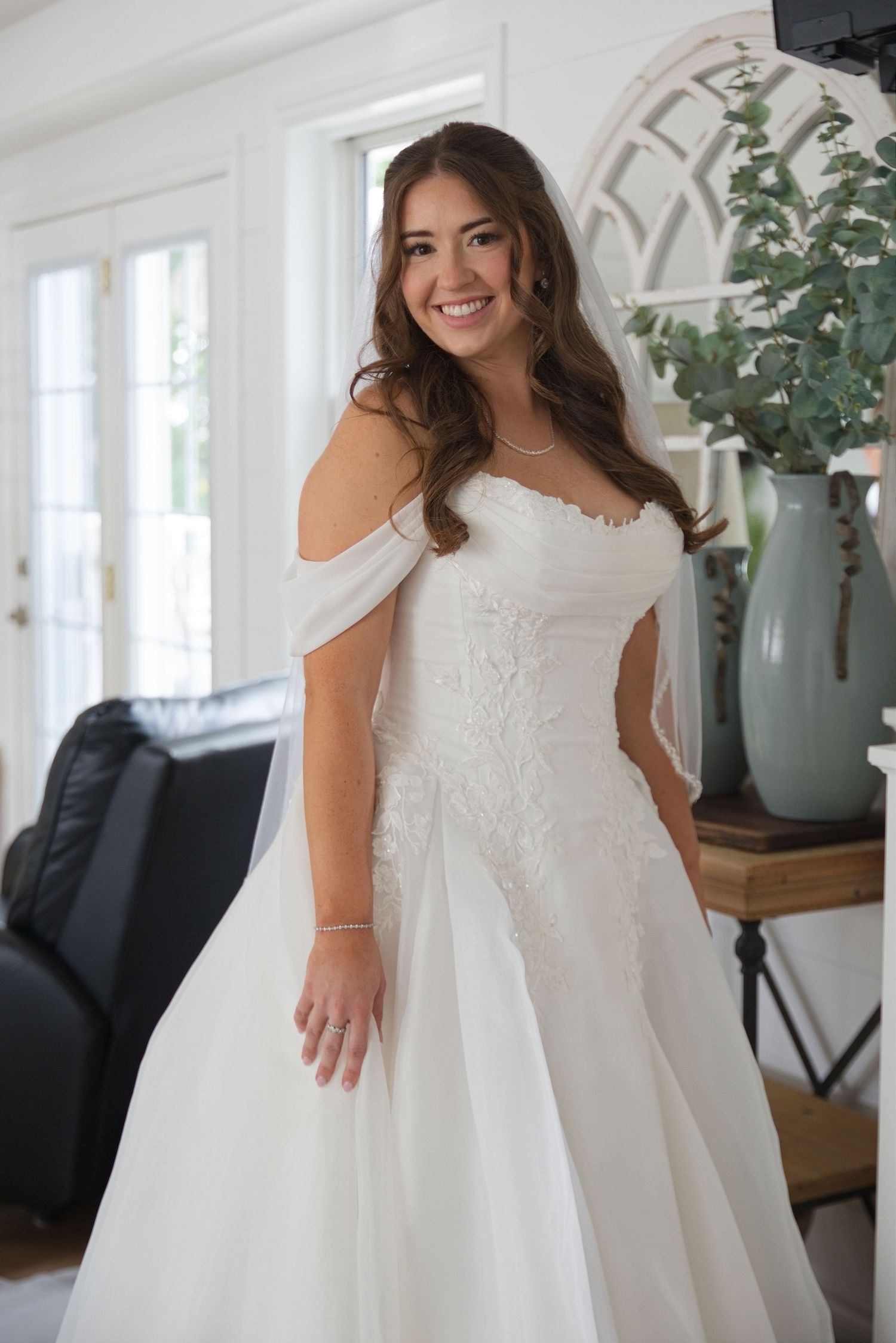 Smiling bride in an off-shoulder white lace wedding gown with a flowing skirt and veil, posing indoors.