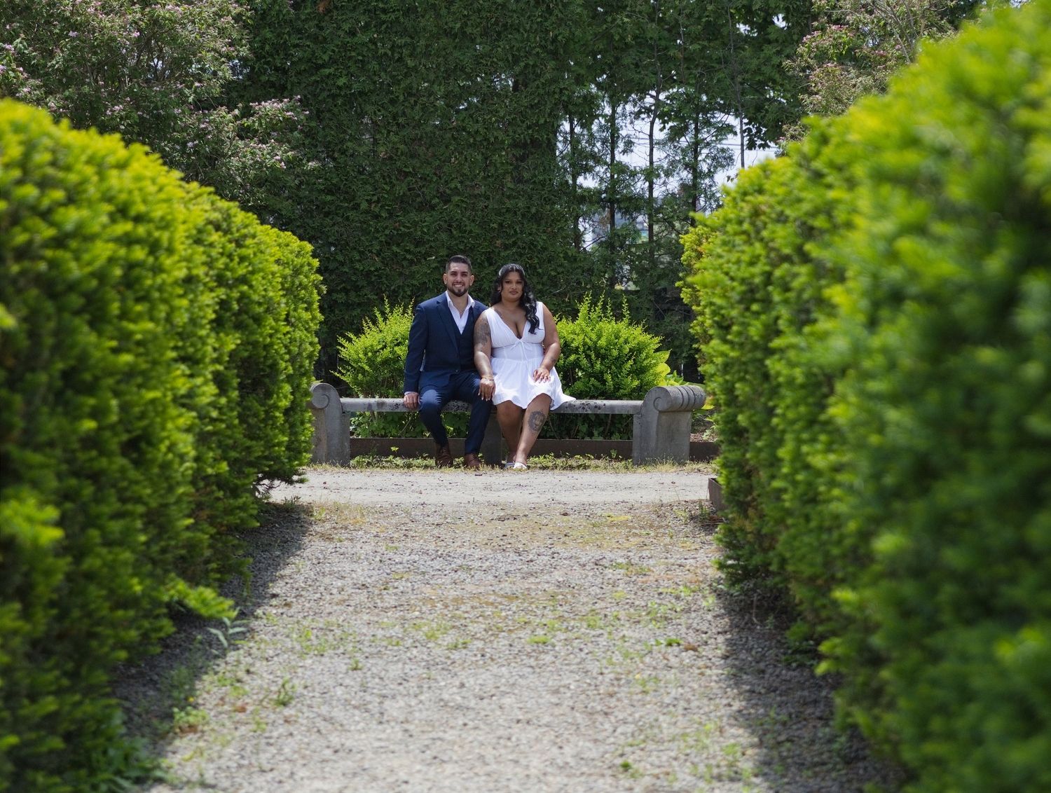 Couple sitting on a garden bench surrounded by manicured hedges and lush greenery on a gravel pathway.