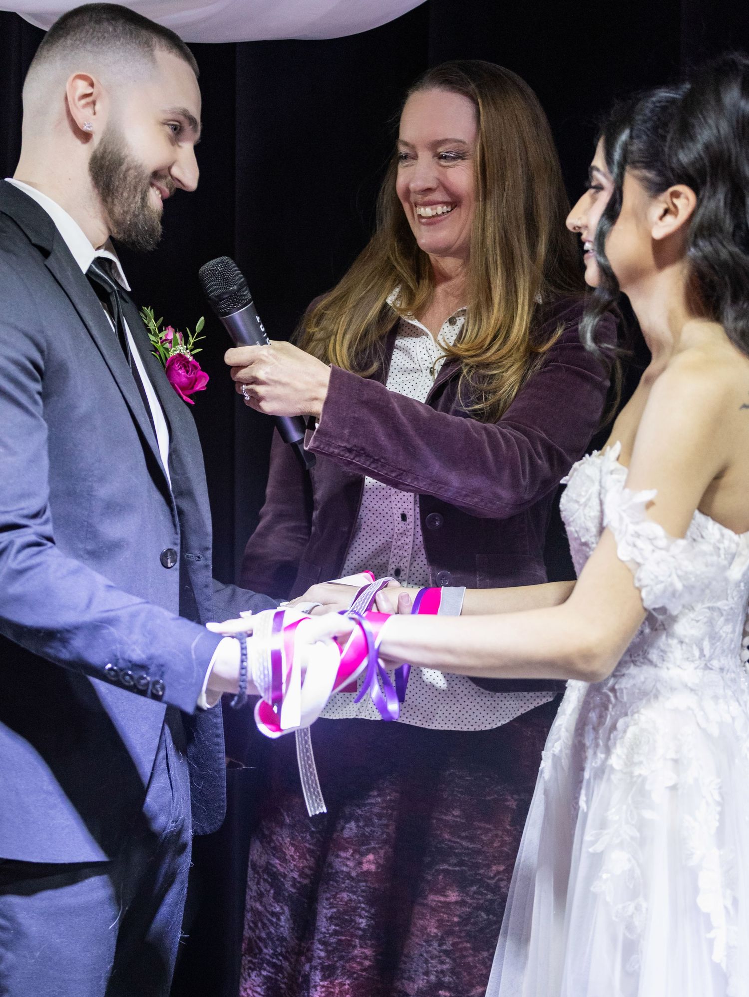 Wedding officiant holding microphone during hand-fasting ribbon ceremony with bride and groom.