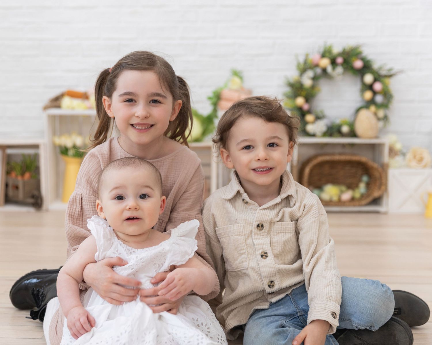 Three siblings posing together with Easter decorations, including a baby in white dress.