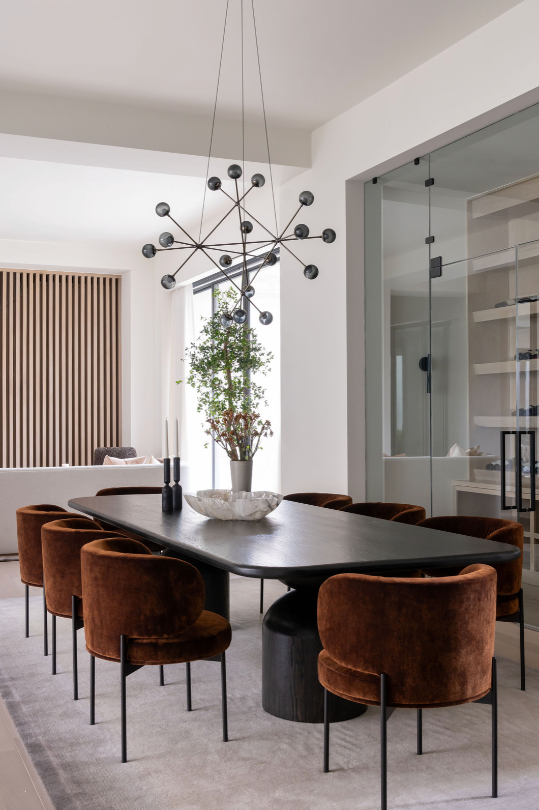 Modern dining room with black oval table, cognac velvet chairs, Sputnik chandelier, and vertical wood slat partition.