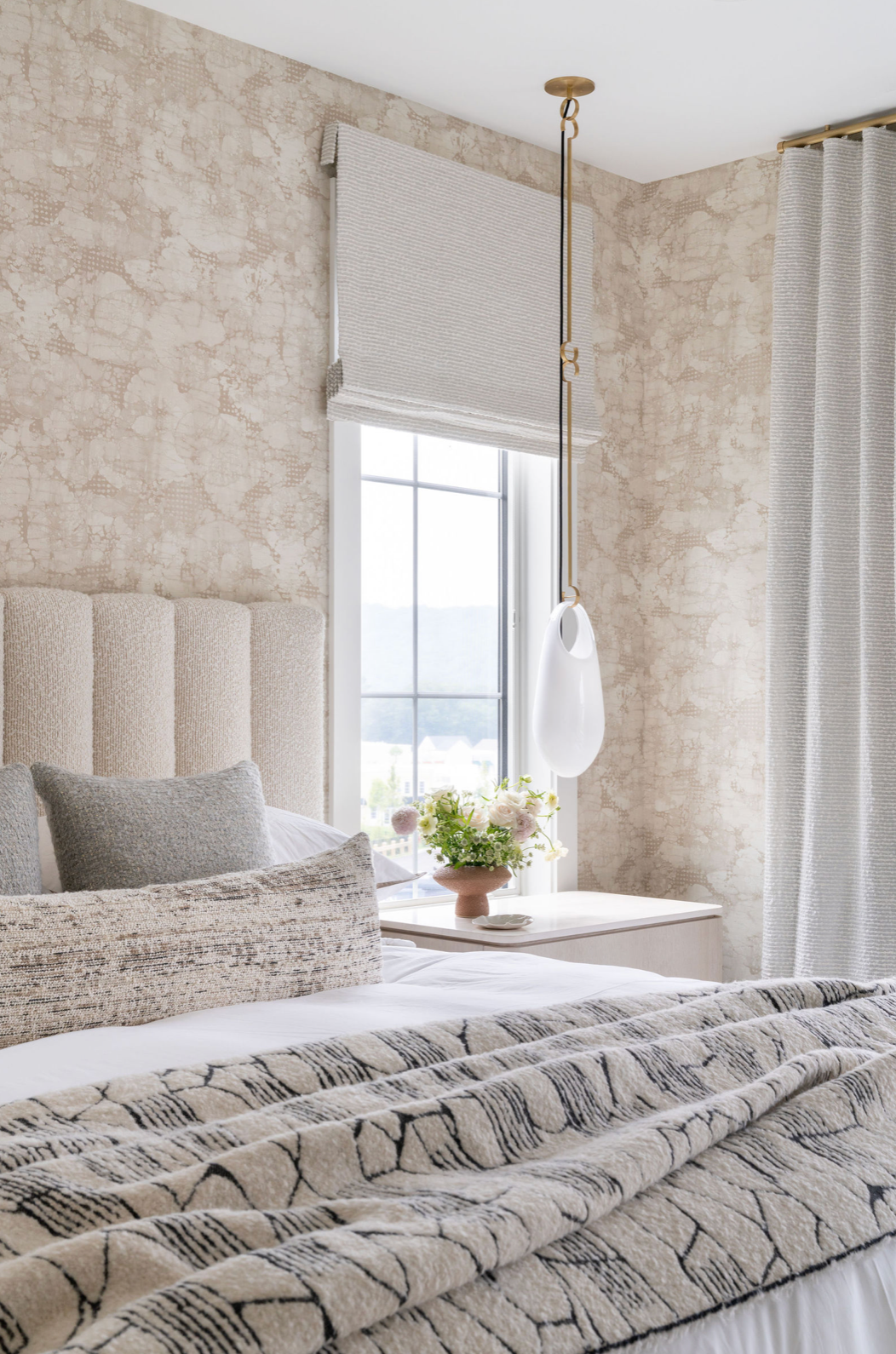 Serene neutral bedroom with textured wallpaper, channeled headboard, patterned throw, and brass pendant light by window.