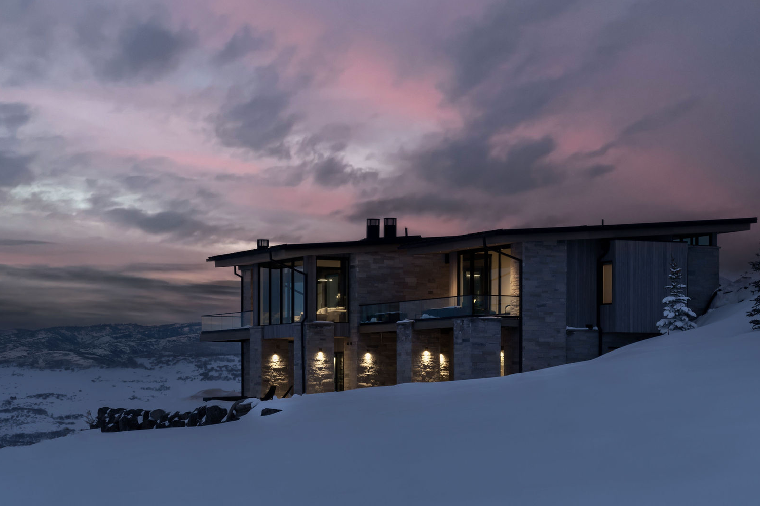 Illuminated luxury mountain home glowing at dusk against a dramatic pink and purple winter sky.