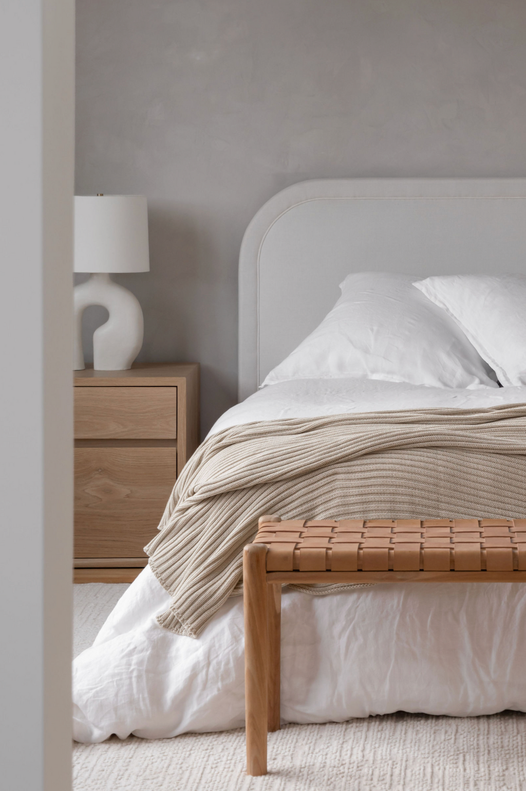 Serene neutral bedroom with arched headboard, linen throw, oak nightstand, and woven leather bench.