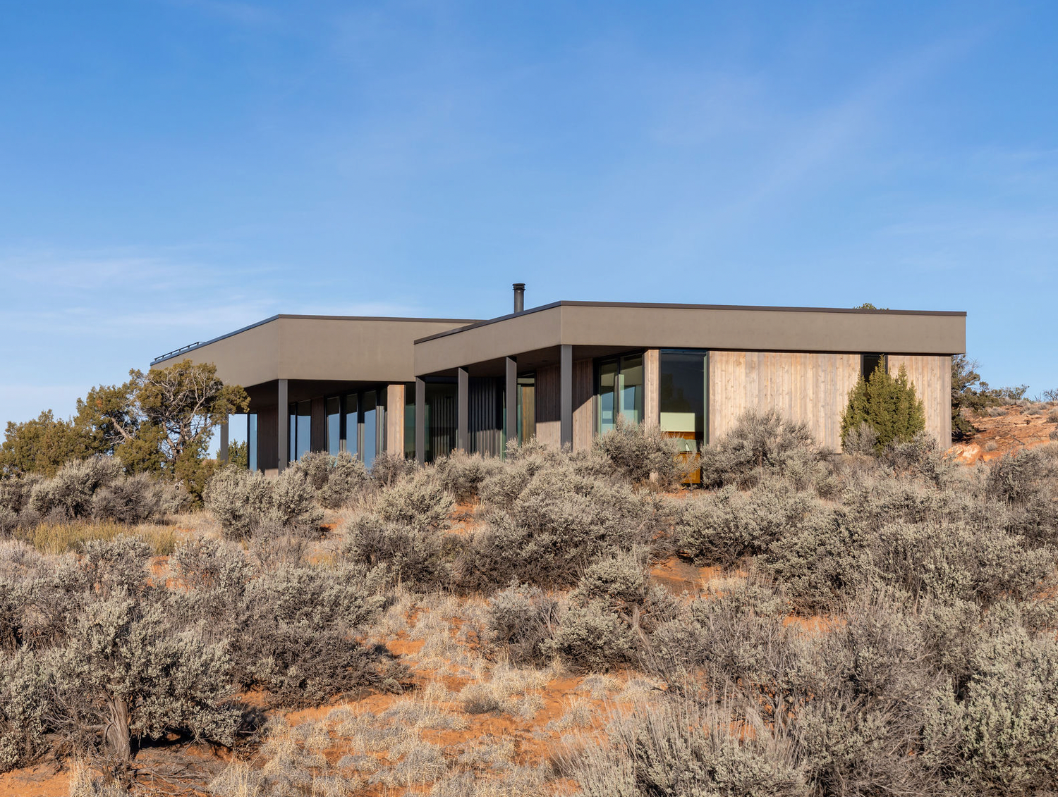 Modern flat-roof desert home with wood siding nestled among sagebrush landscape.