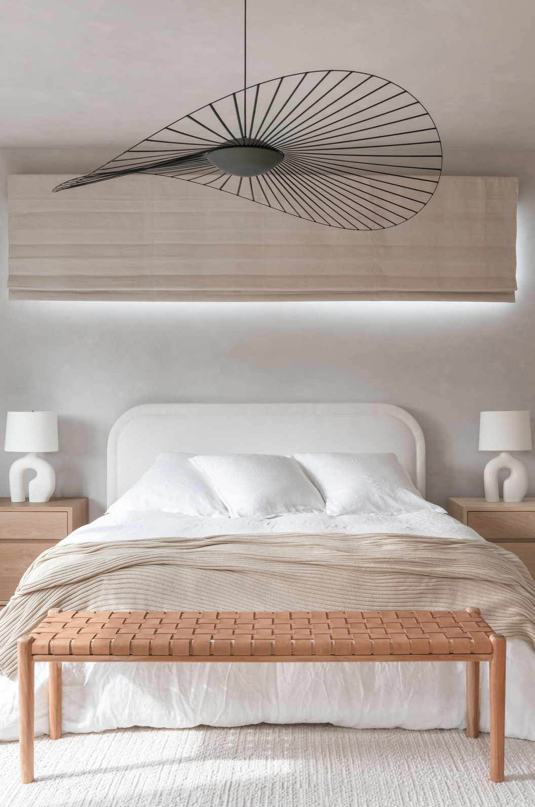 Minimalist bedroom with white upholstered bed, beige throw, woven bench, and black Vertigo pendant light.