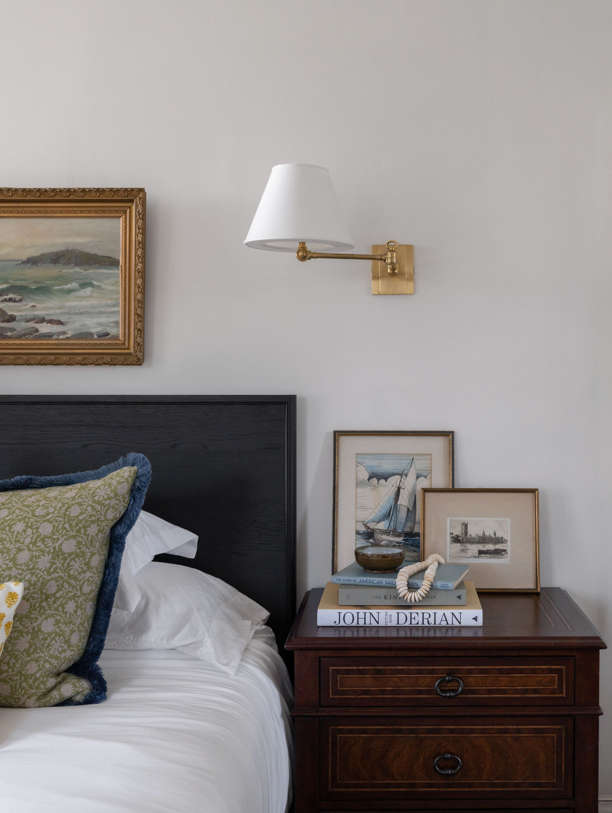 Bedroom nightstand with stacked books, framed coastal artwork, brass swing-arm wall sconce, and navy upholstered headboard.
